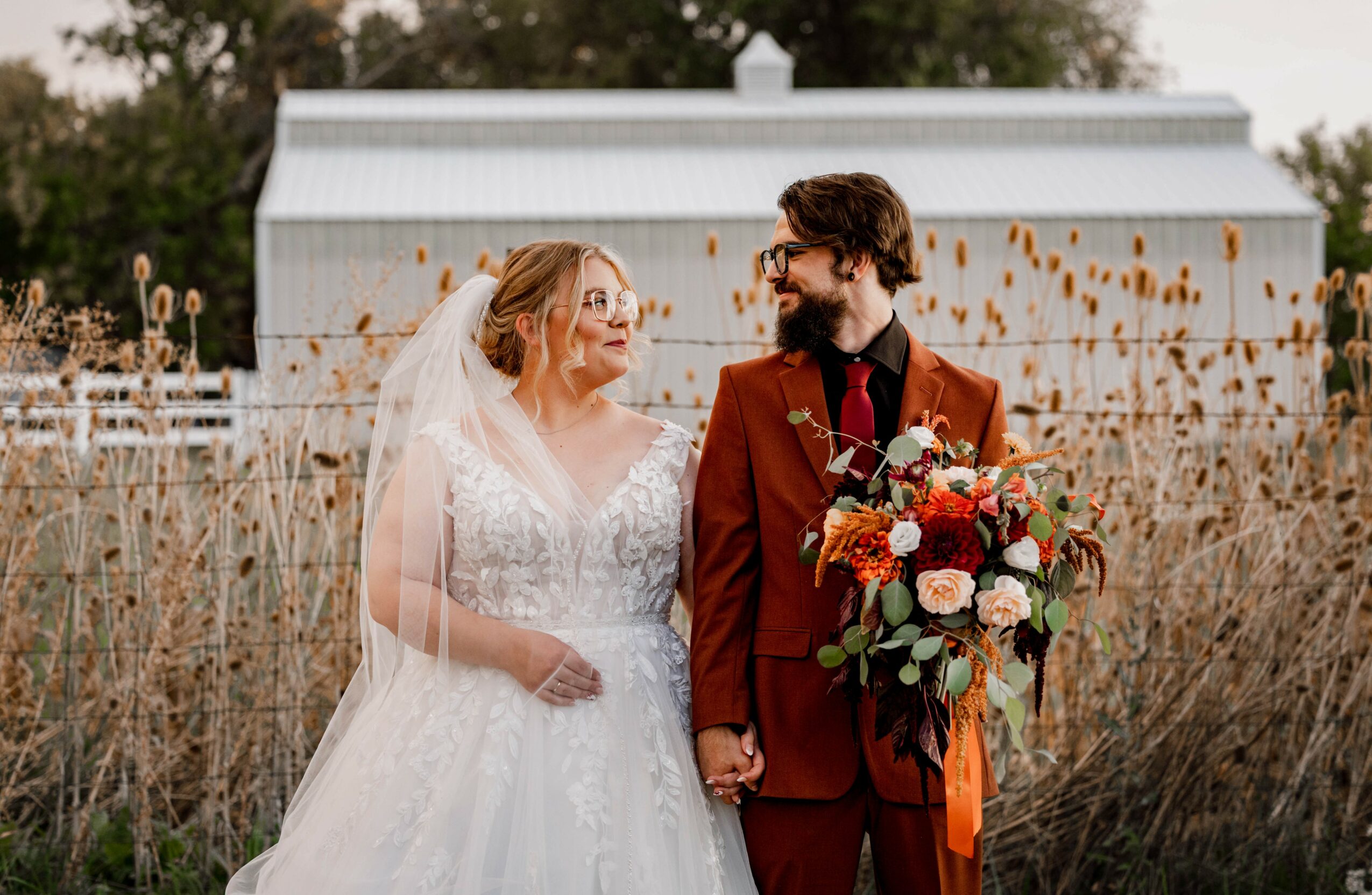 Bride and groom holding hands outdoors, bride in white wedding dress, groom in red suit, with a bouquet, in front of a white building.