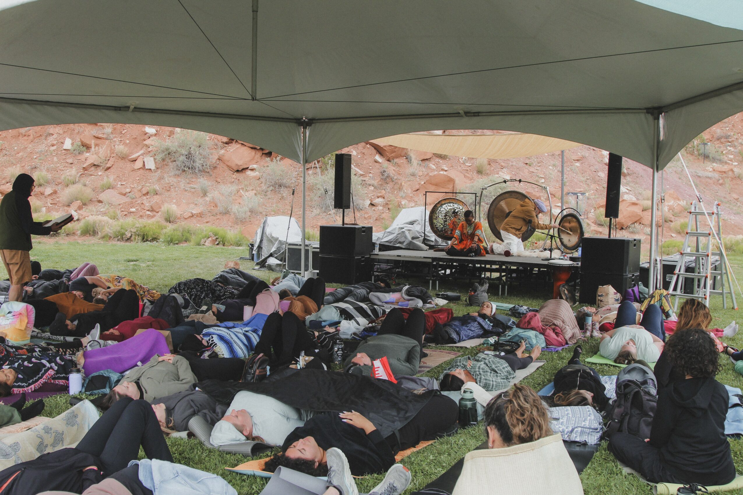 People lying on grass under a tent, facing a stage with speakers and equipment outdoors.