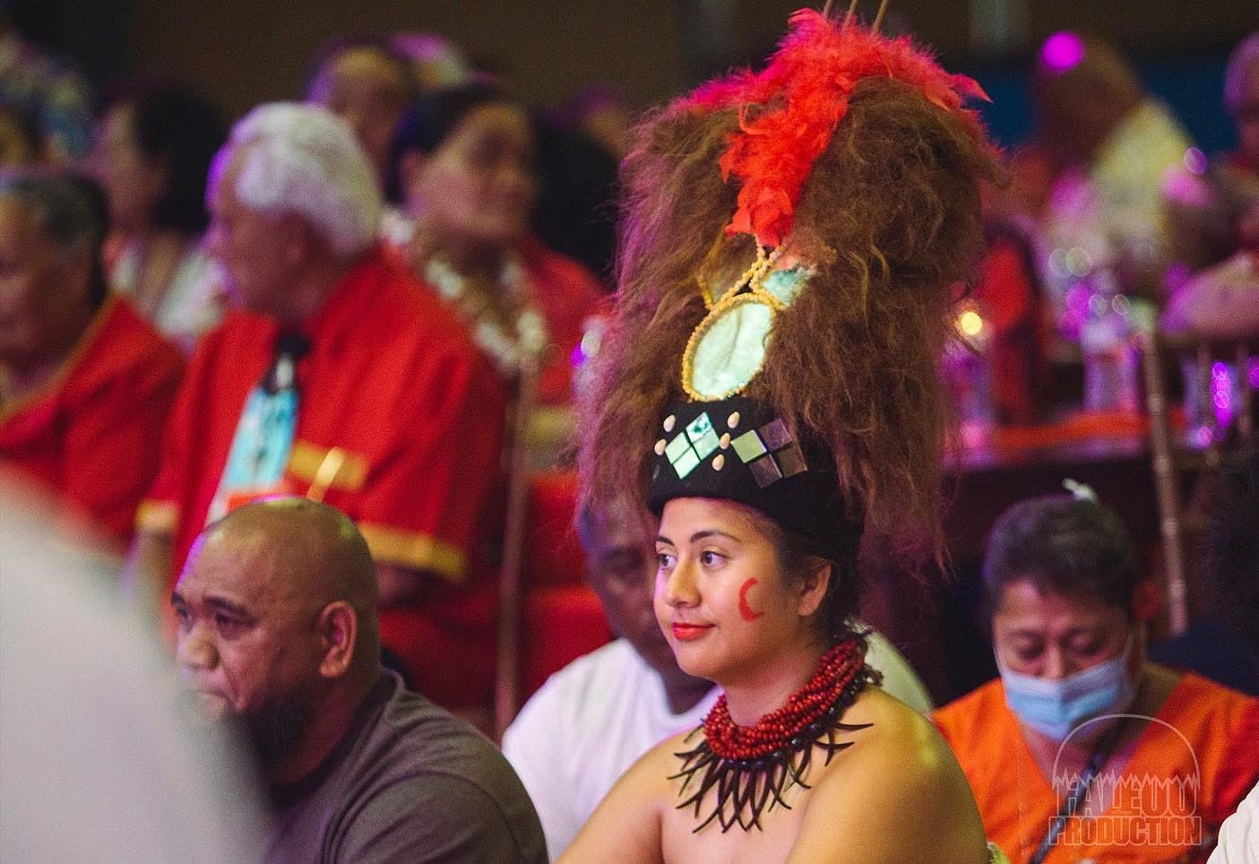 Woman in traditional costume with large feathered headdress, seated among other people at an indoor event.