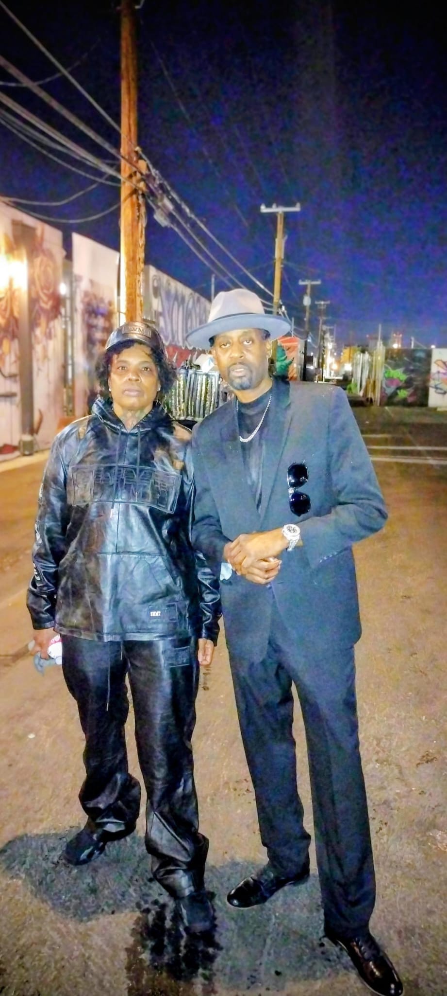 Two people stand on a street at night, wearing dark clothing, with streetlights and utility poles in the background.