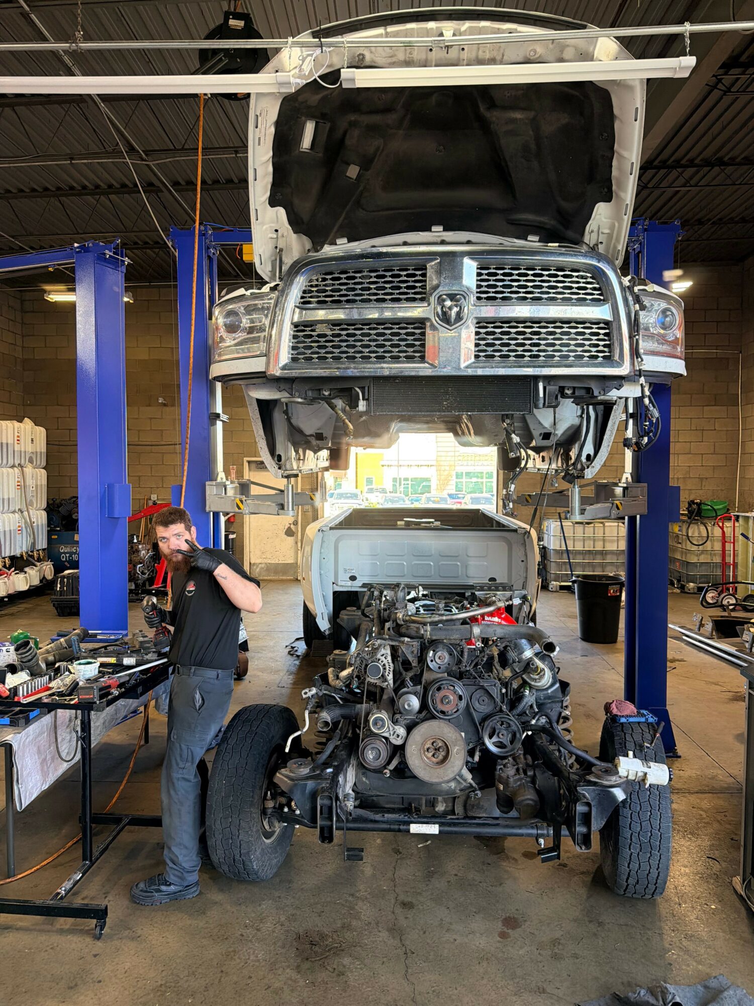 Person standing next to a large vehicle engine in a workshop with a vehicle lifted above on a blue lift.