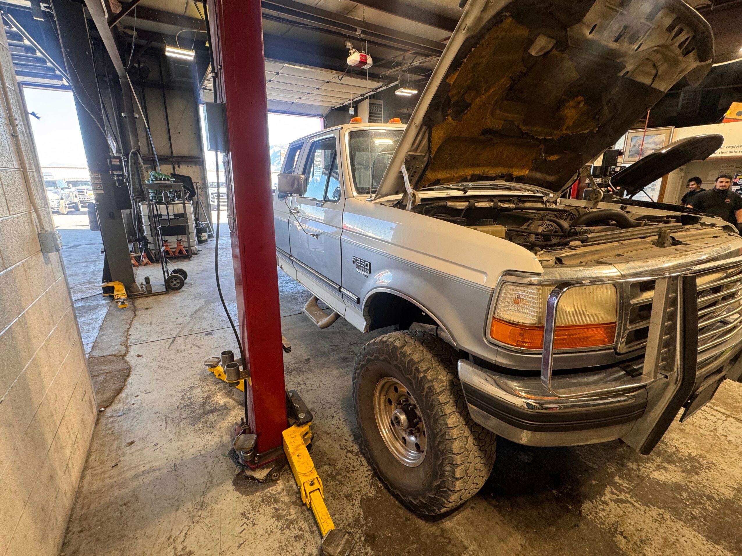 White pickup truck with open hood inside garage, vehicle lifted on a hydraulic lift, tools and equipment nearby.