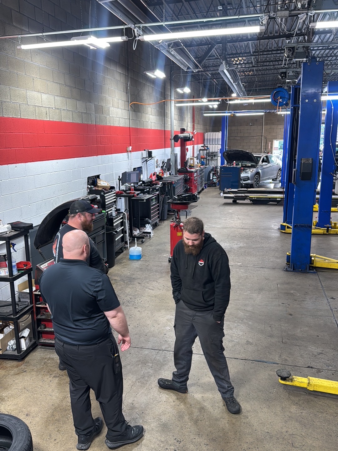Three men standing and talking in an automotive workshop with tools and cars in the background.
