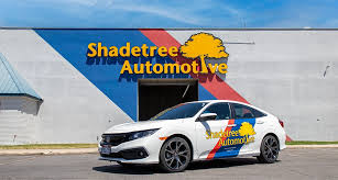 White car with colorful logo parked in front of a building with a sign reading Shadetree Automotive.