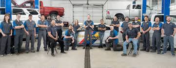Group of people standing and sitting in a garage with cars on lifts in the background.