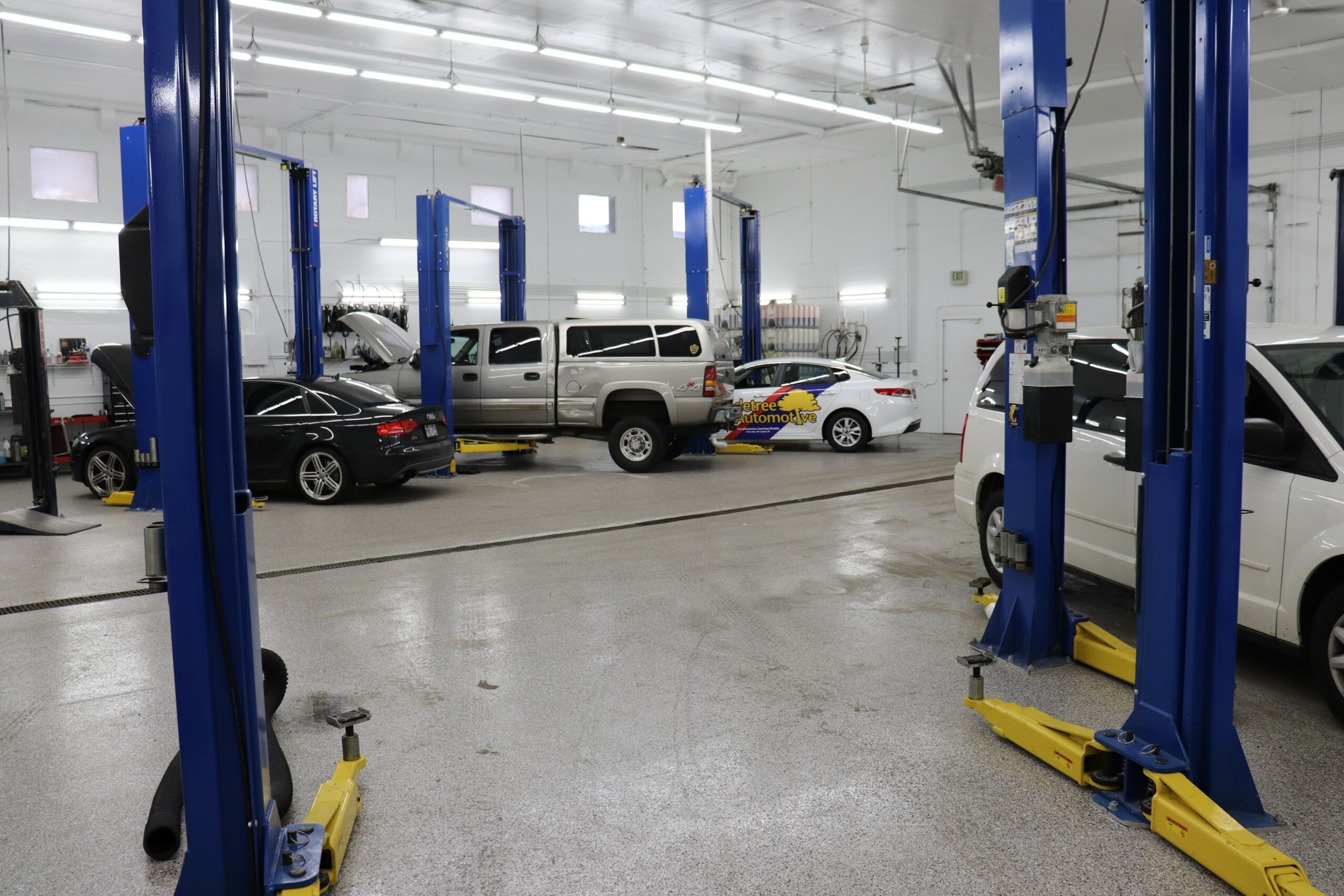 Indoor car repair shop with vehicles on lifts and tools, bright lighting, and white walls.