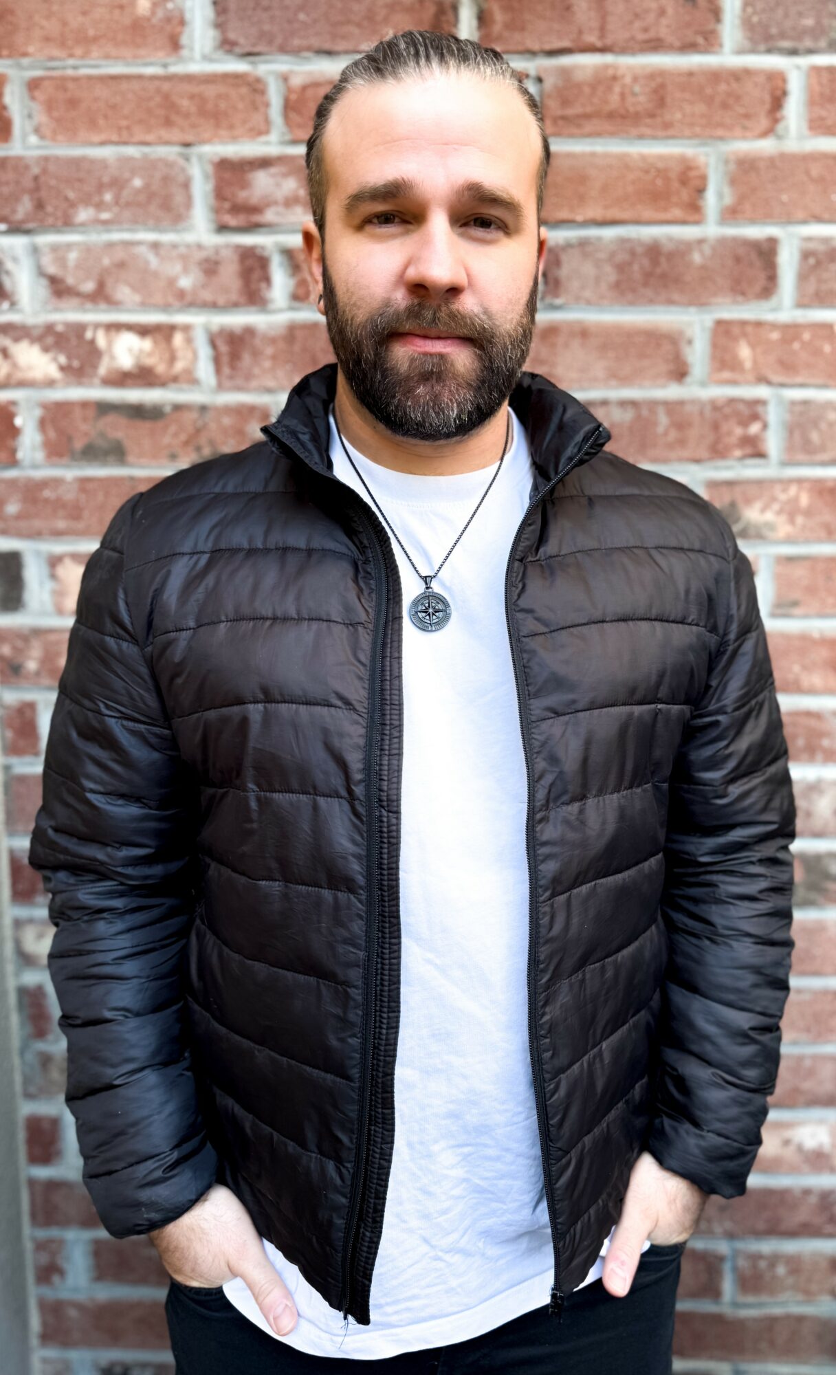 Man with beard and short hair wearing a black puffer jacket and white shirt, standing against a brick wall.