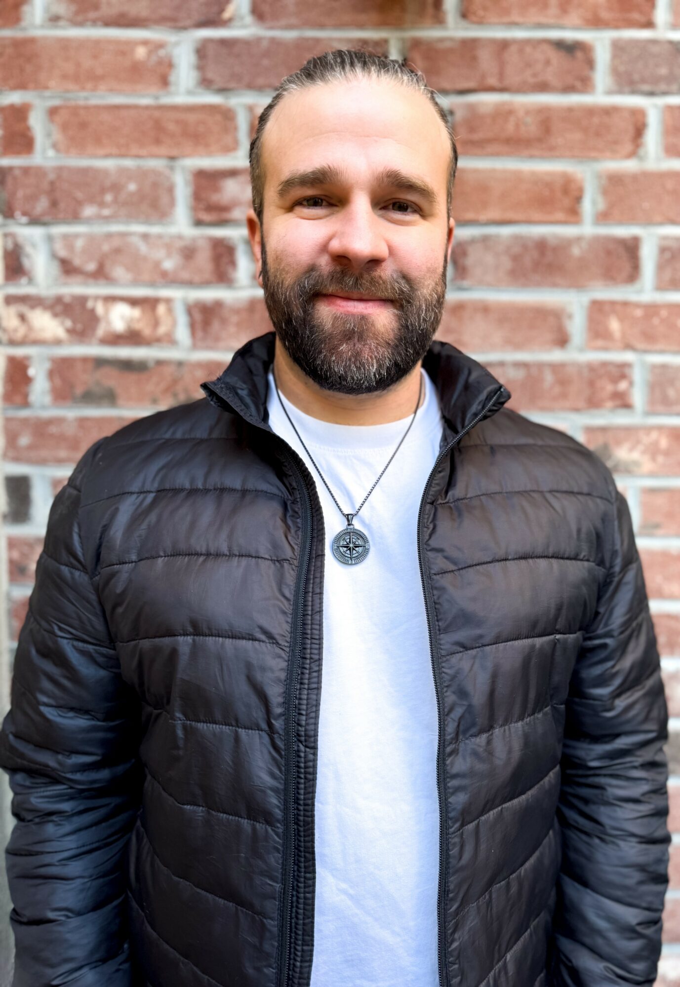 Man with beard and mustache wearing a black puffer jacket and a necklace with a pendant, standing in front of a brick wall.