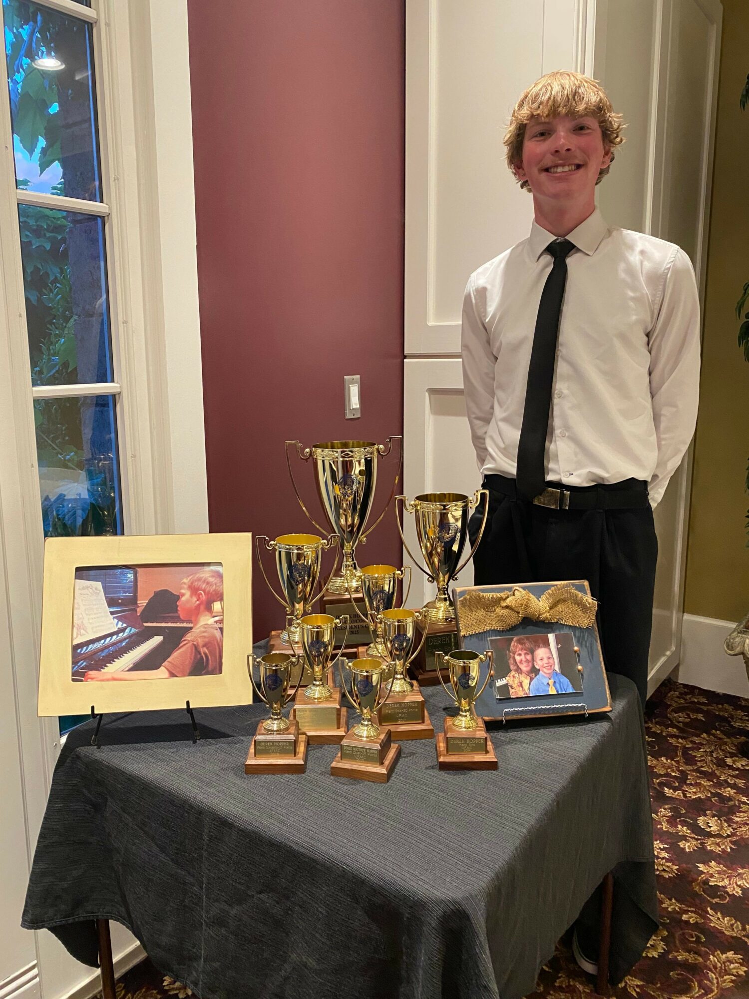 Young man in white shirt and black tie standing next to a table with trophies and framed photos.