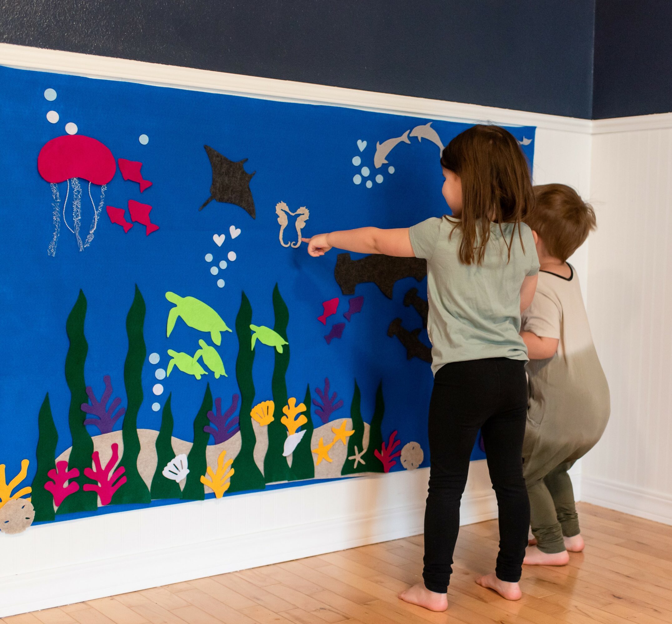 Two children stand in front of an underwater-themed wall display with sea creatures and plants, pointing at it.