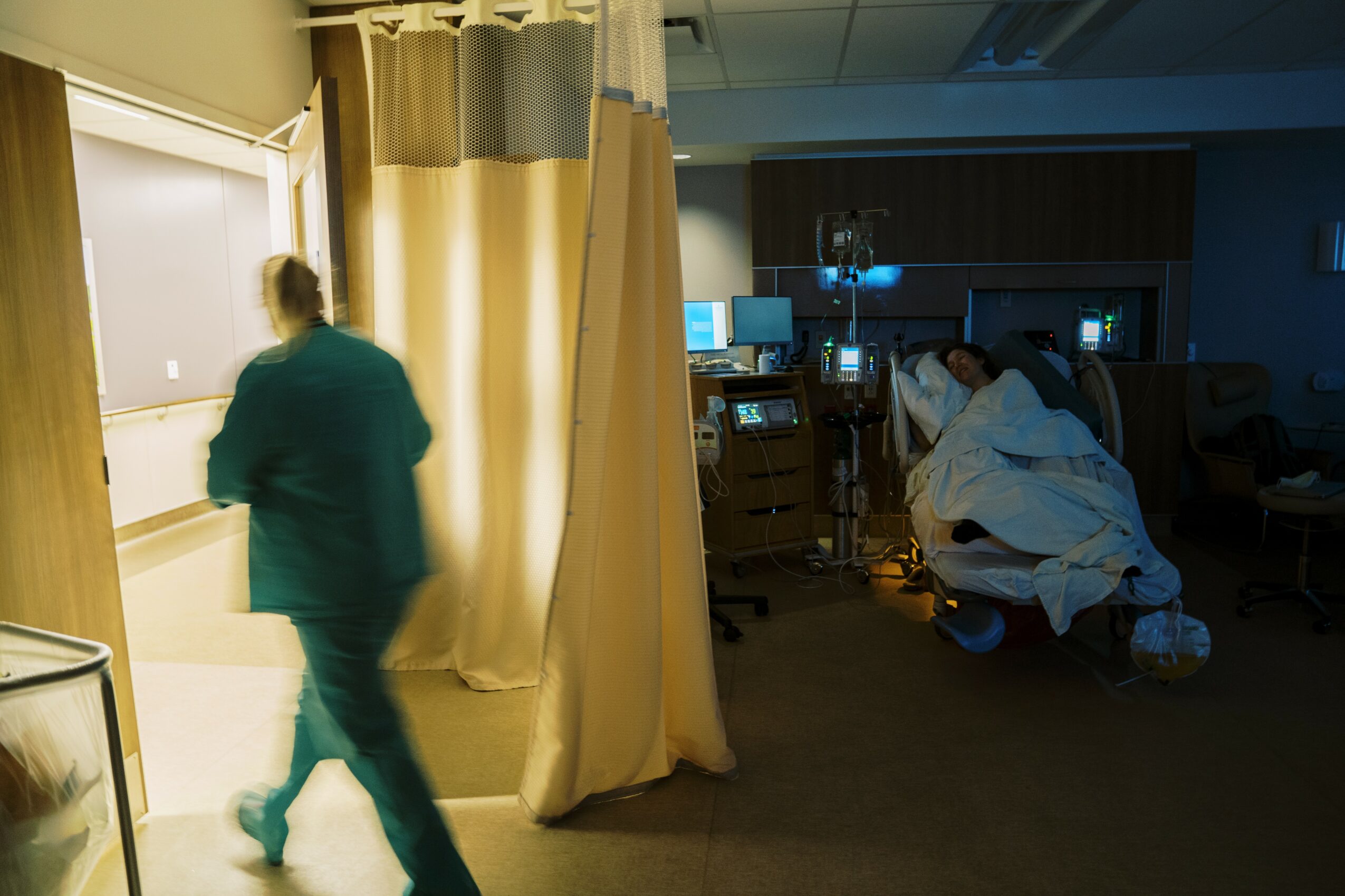 Hospital room with a person in scrubs walking past a curtain, and medical staff attending to a patient on a bed.