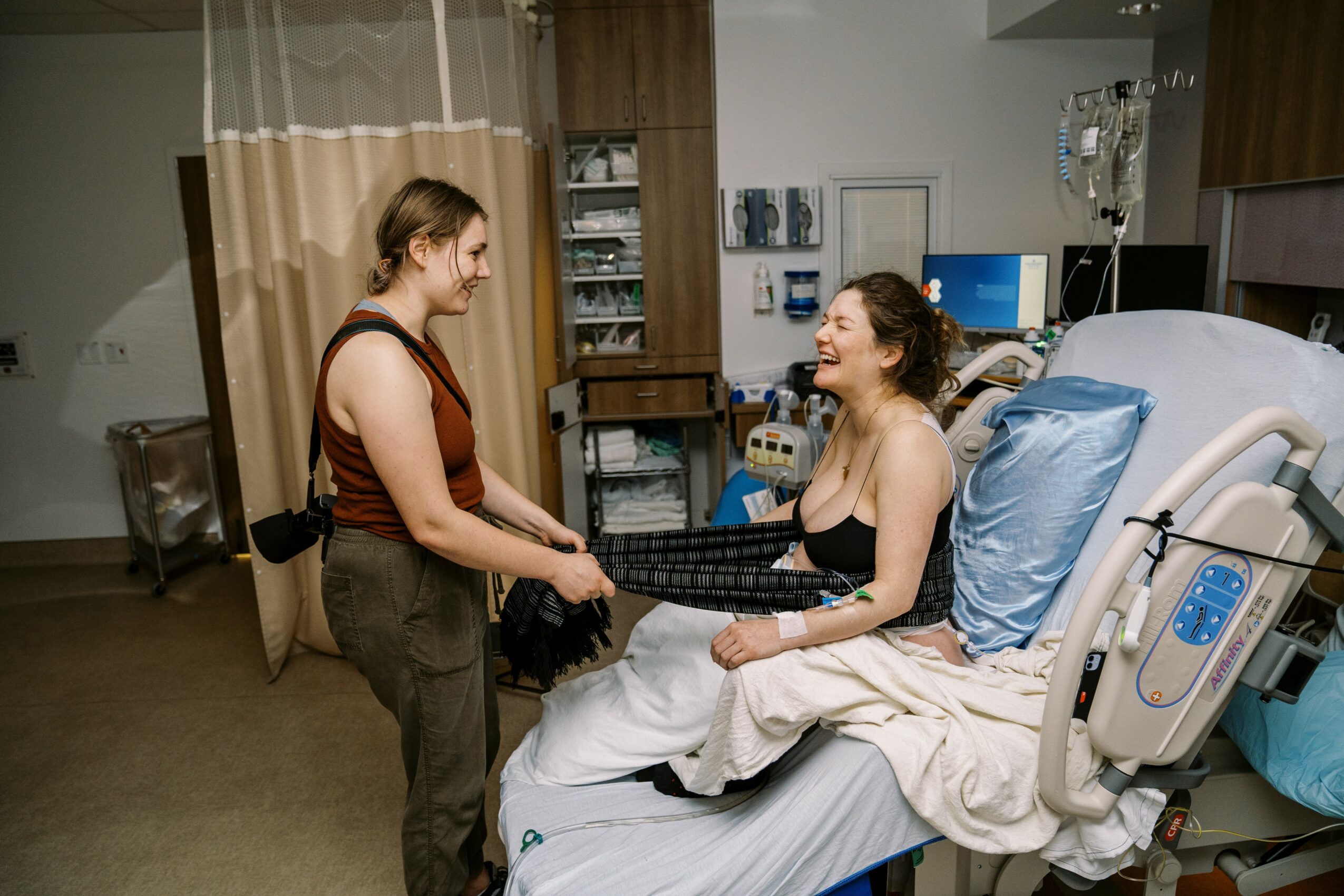 Two women in a hospital room, one sitting on a bed and the other standing, smiling and holding hands.