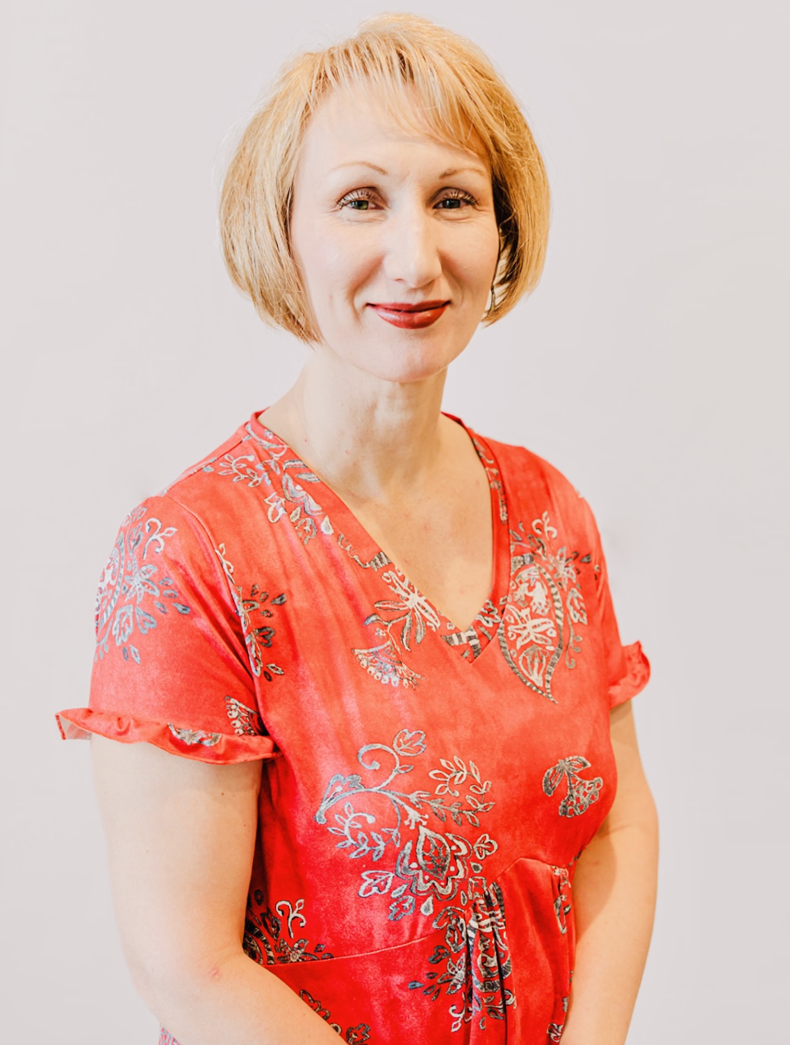 Woman with short blonde hair wearing a red patterned dress, smiling, against a plain background.
