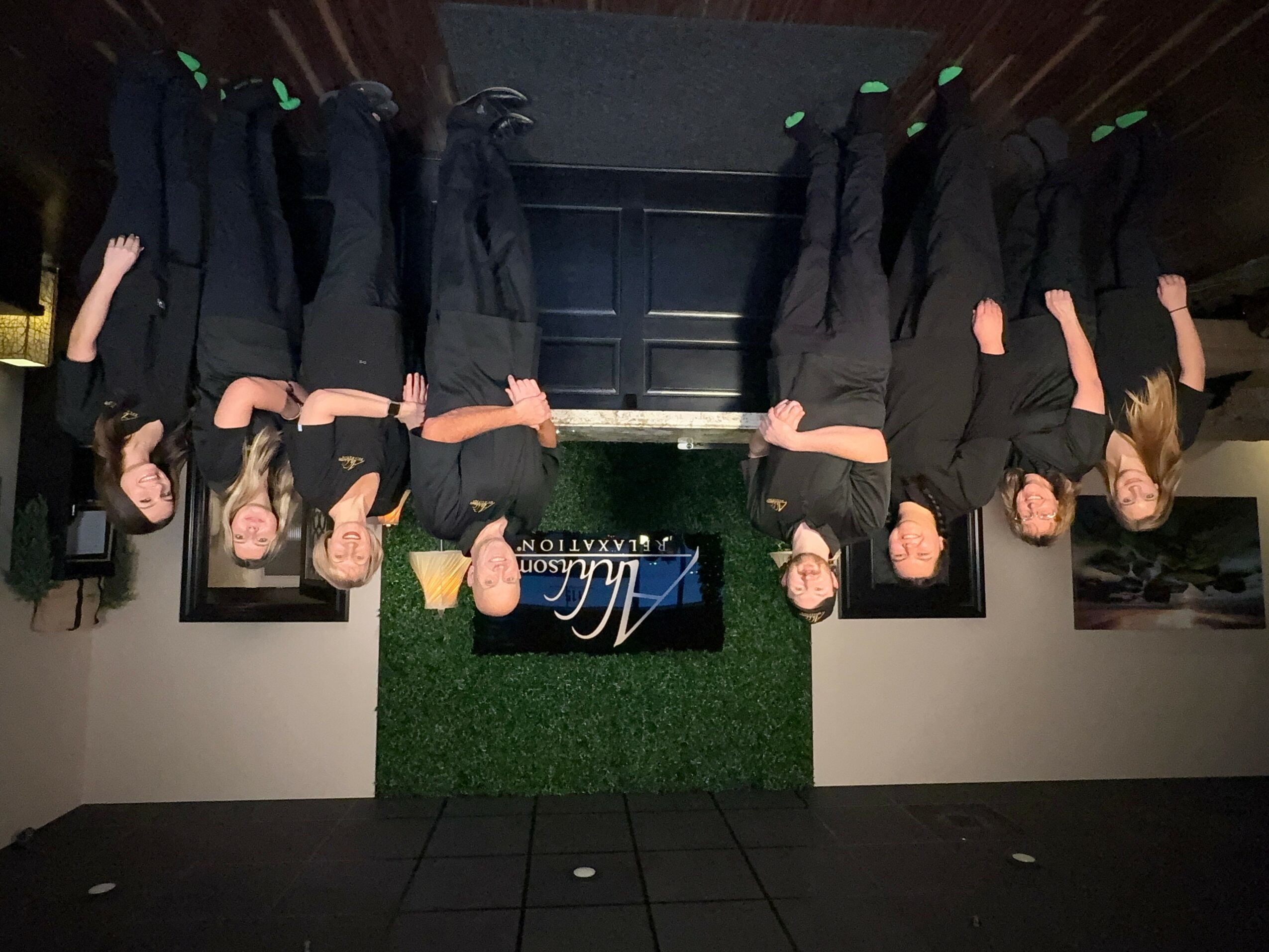 Group of eight people standing indoors, some with arms raised, in front of a green wall with a sign.