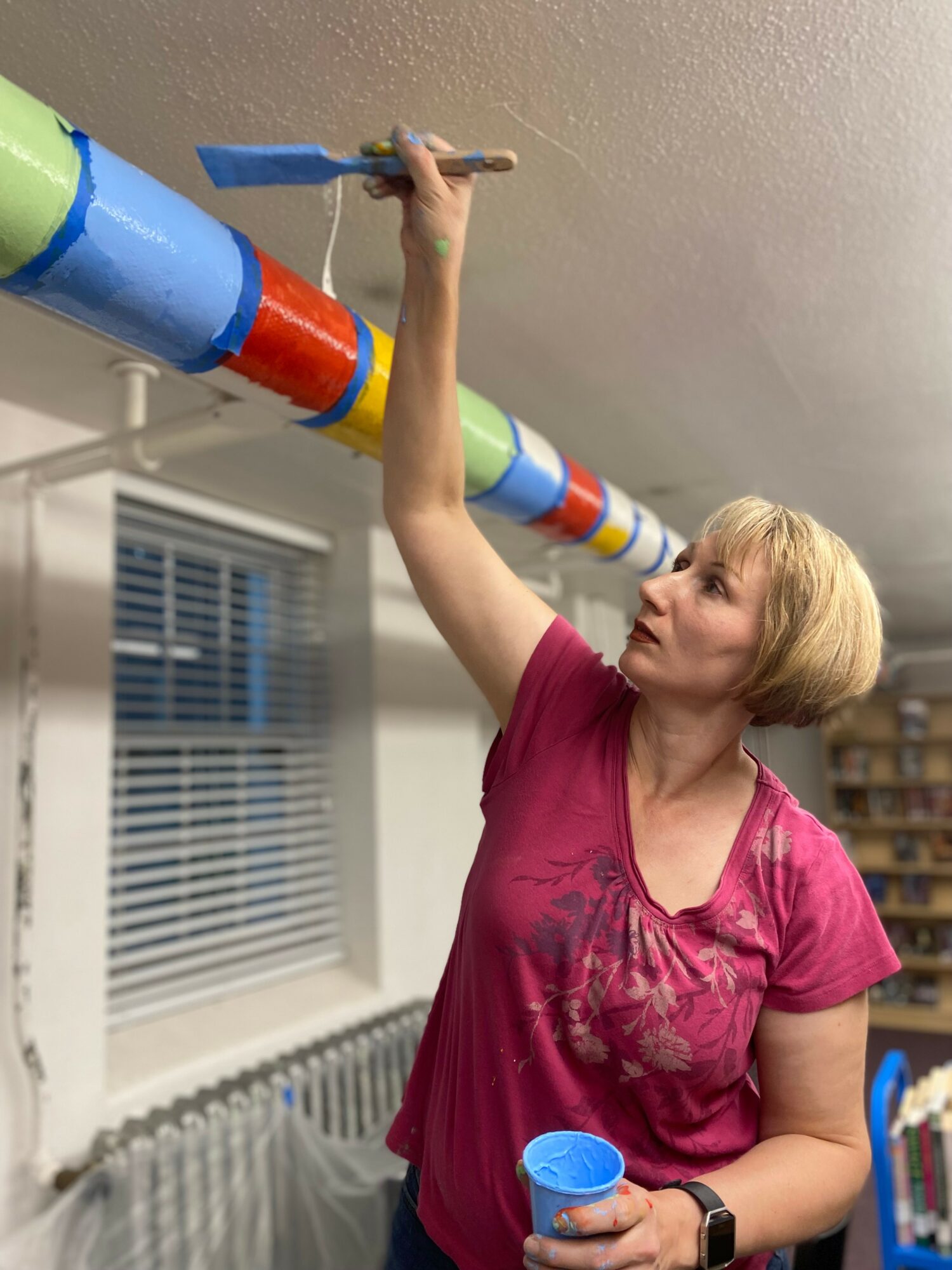 Woman painting a colorful pipe on ceiling indoors, holding a paintbrush and a small container, wearing a pink shirt.