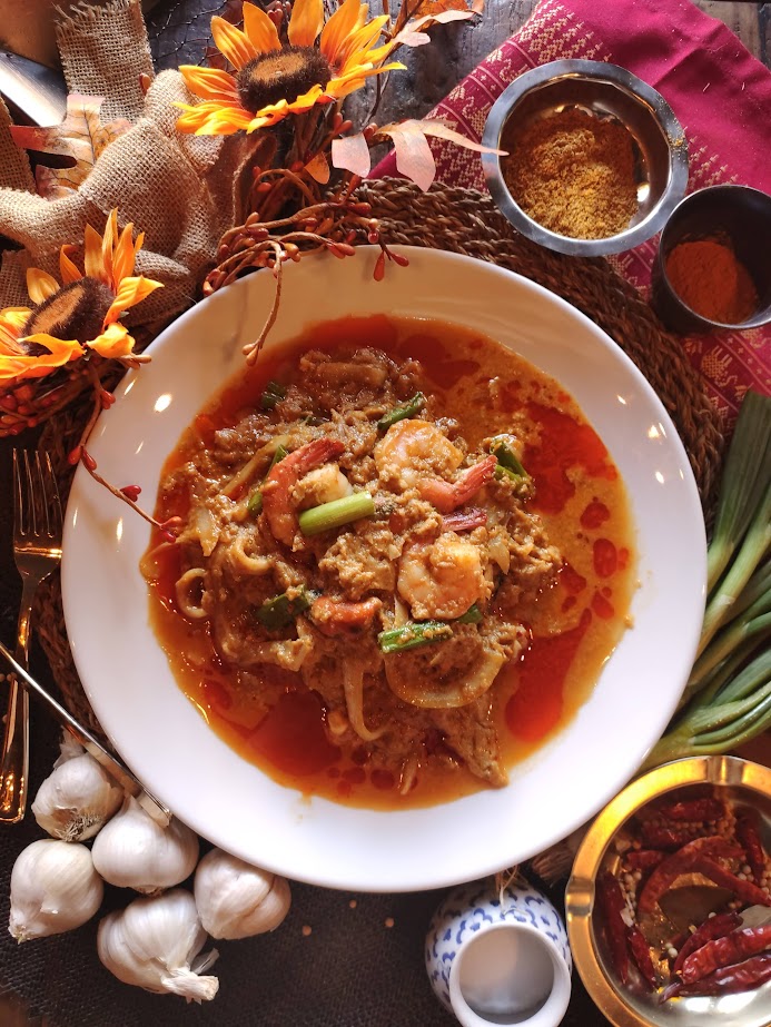 Bowl of spicy noodle soup with vegetables and meat, surrounded by garlic, sunflower flowers, and condiments on a table.