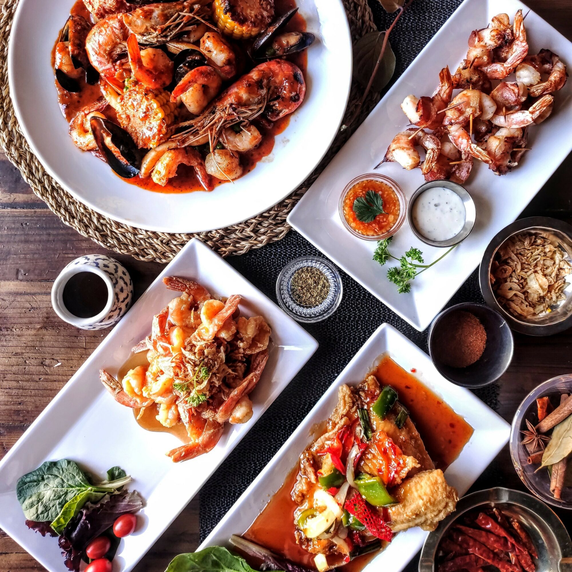 Assorted dishes of seafood, vegetables, and sauces on a dark table with bowls and garnishes.