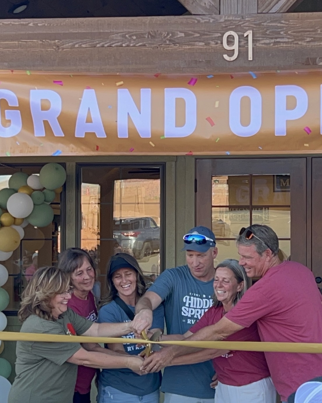 Group of five people holding a ribbon outside a store with balloons and a grand opening sign.