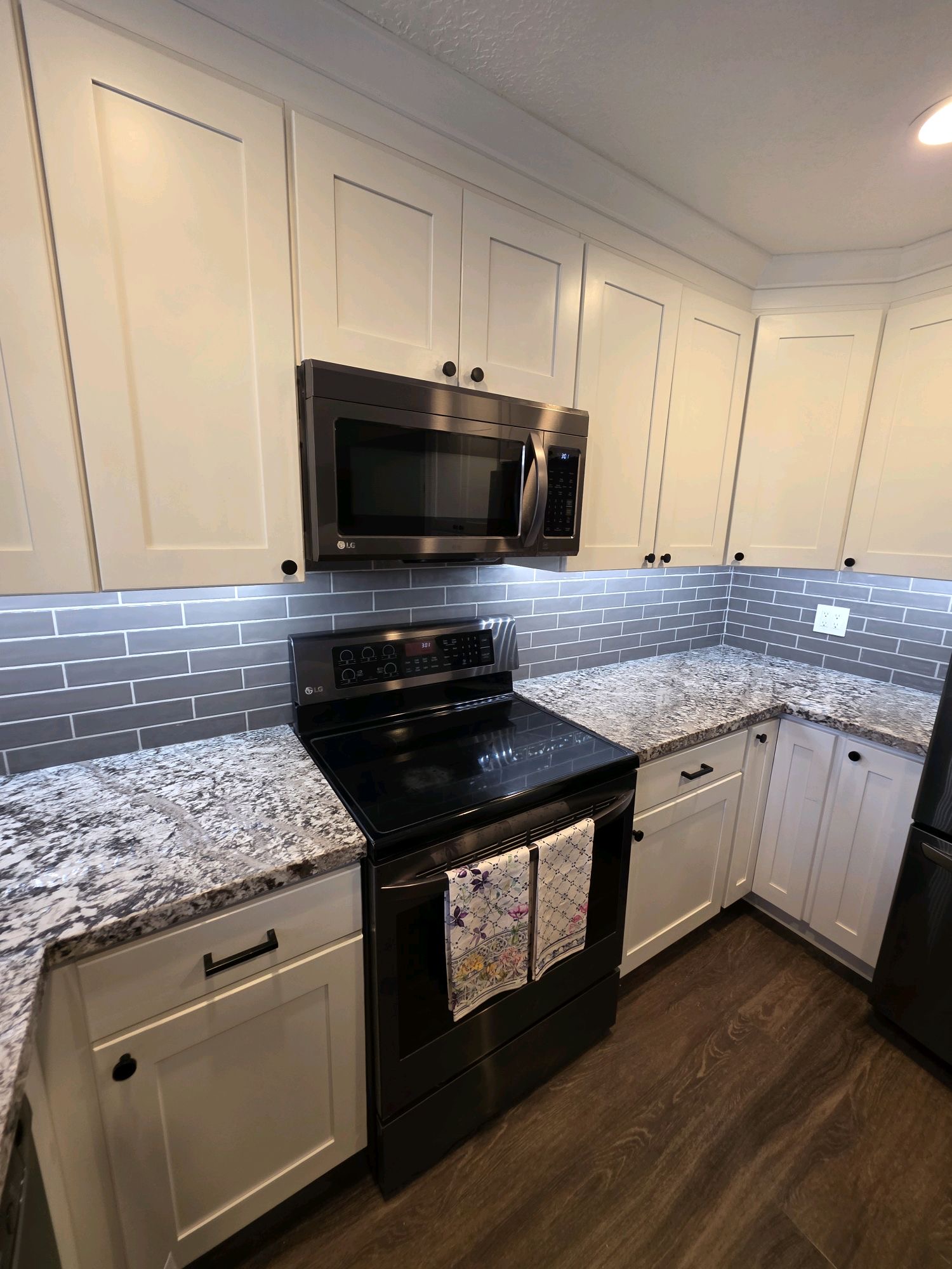 Kitchen with white cabinets, granite countertop, gray backsplash, black stove, microwave, and hardwood floor.