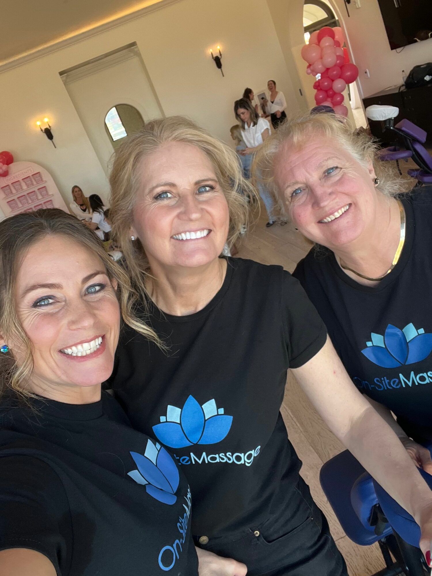 Three women smiling in a room with balloons and people in the background, wearing matching black shirts with a blue lotus logo.