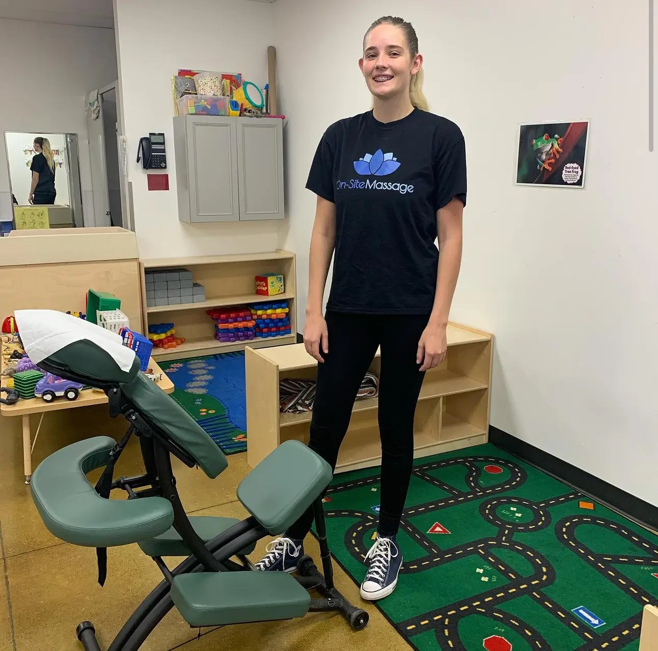 Young woman standing in a room with a massage chair, shelves, and a colorful carpet, smiling at the camera.