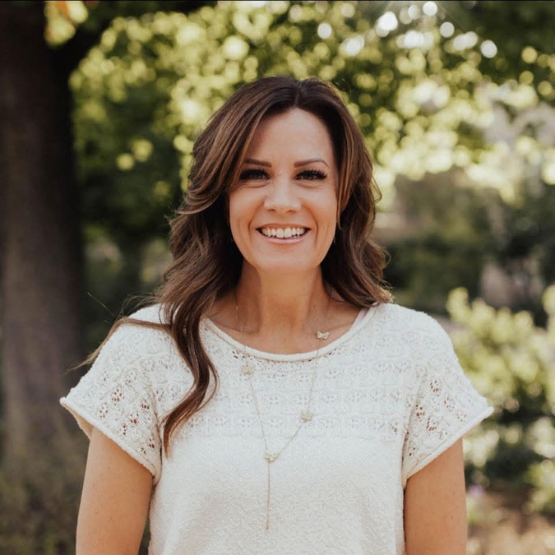 Smiling woman with wavy brown hair outdoors, wearing a white lace top, trees and sunlight in background.