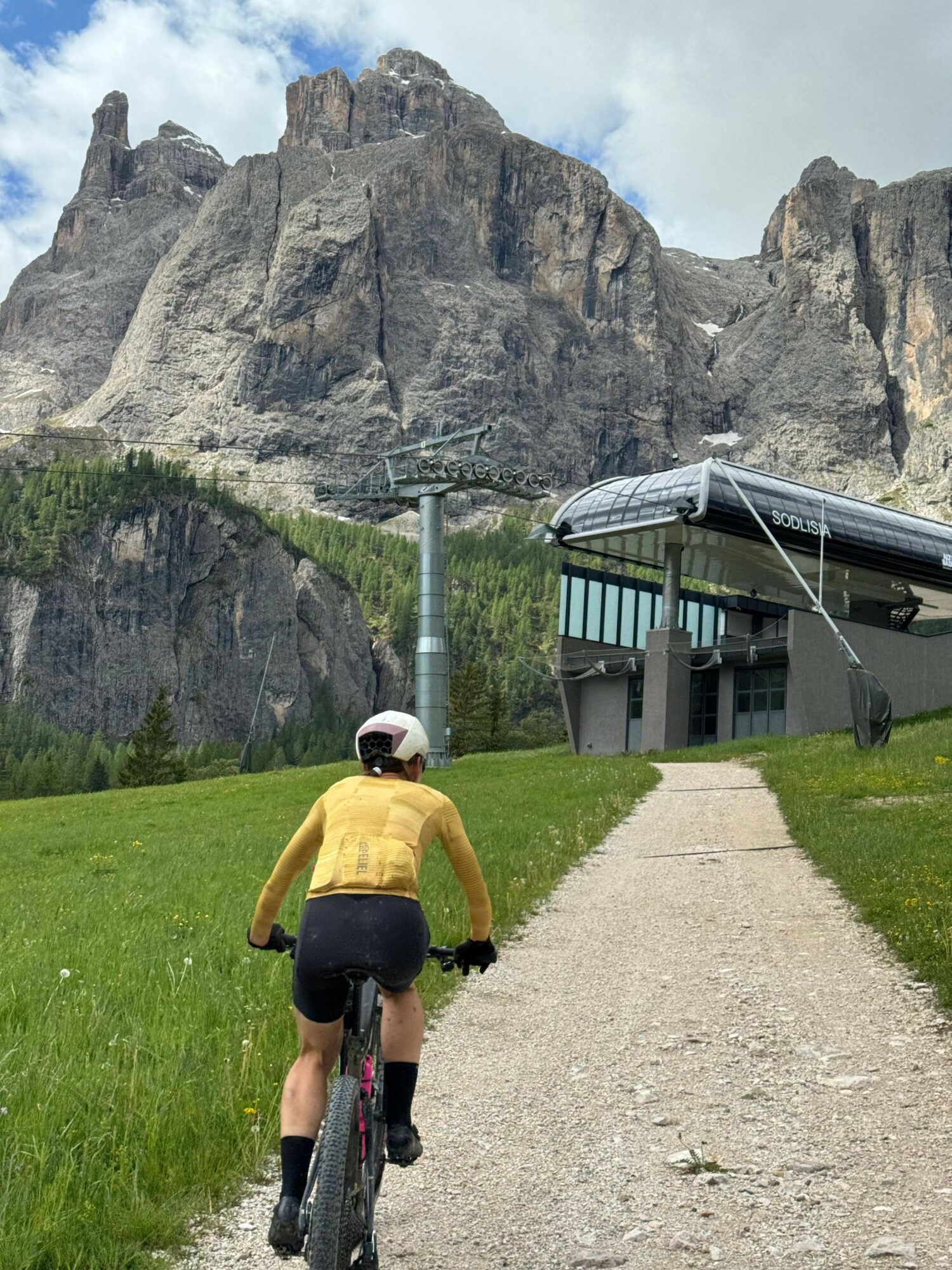 Person riding a bicycle on a grassy path near rocky cliffs and a building with a metal roof.