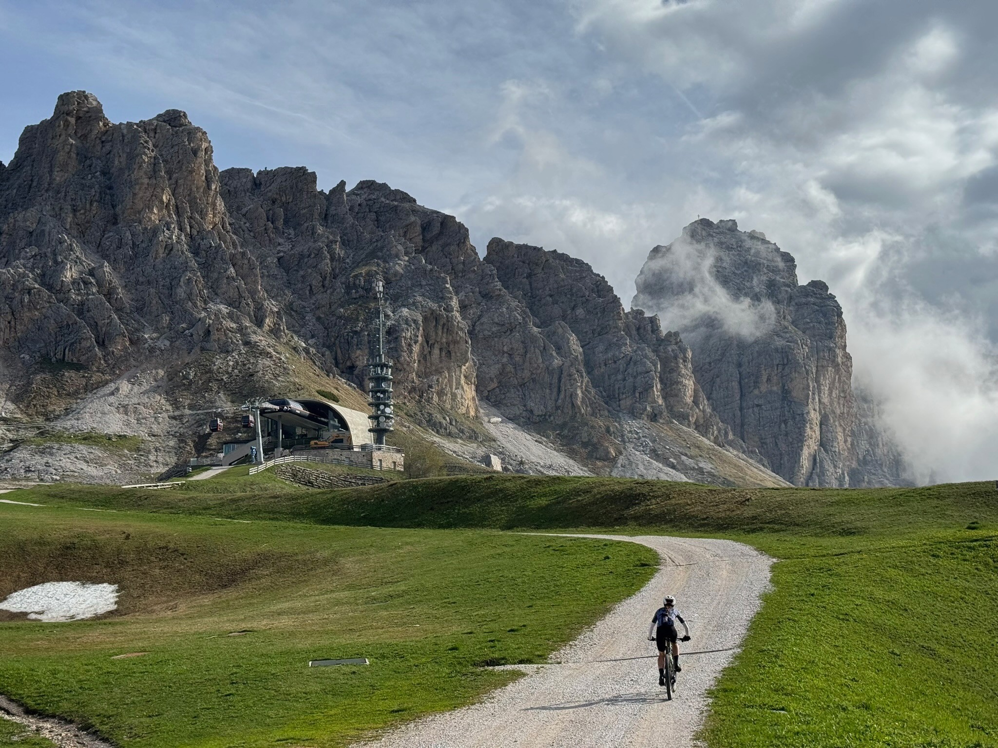 Person walking on a gravel path through green grass with mountain cliffs in the background and cloudy sky.