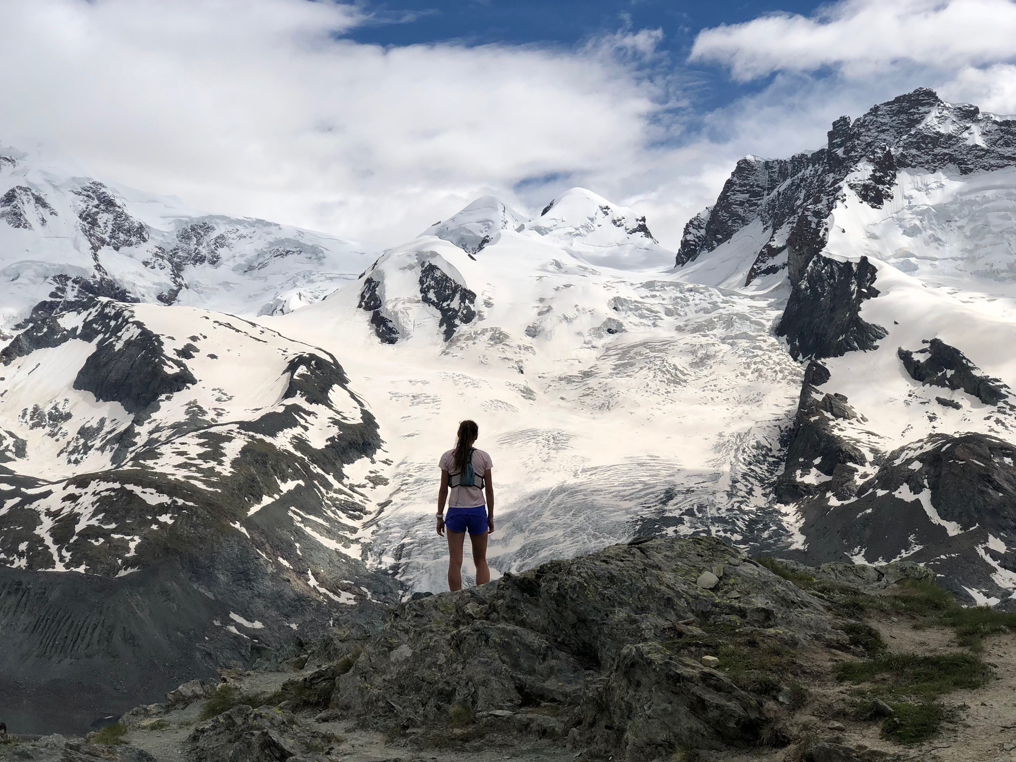 Person standing on rocky terrain facing snow-covered mountains and cloudy sky, with some dark rocks and patches of grass.