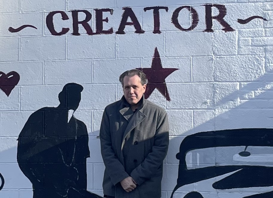 Man standing in front of a wall with the word 'CREATOR' and star, shadow cast on wall, bright sunlight.
