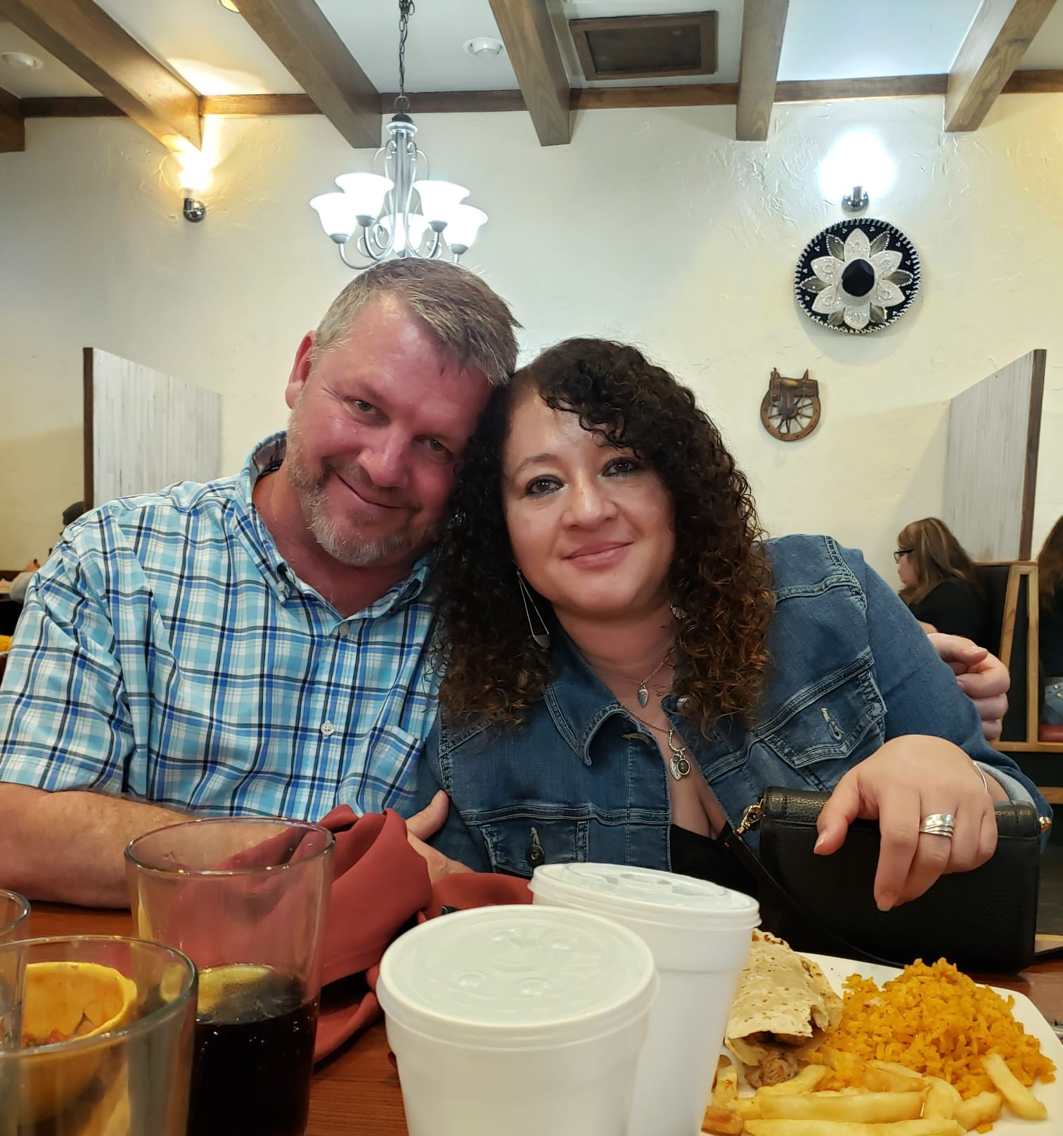 Two people sitting at a table, smiling, with food and drinks, in a decorated indoor setting.