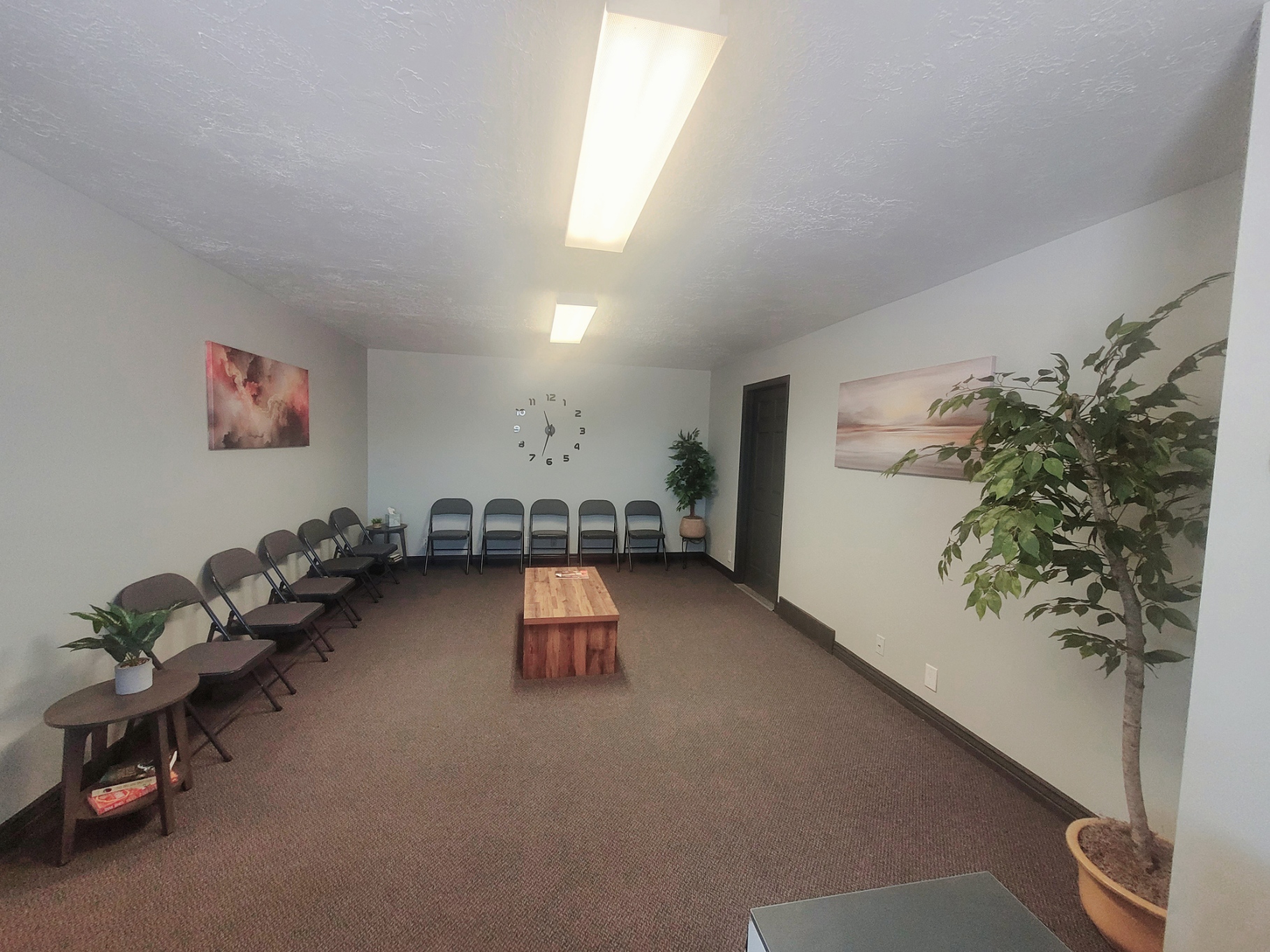 Empty room with chairs along walls, a small wooden table, potted plants, and wall art, lit by ceiling lights.