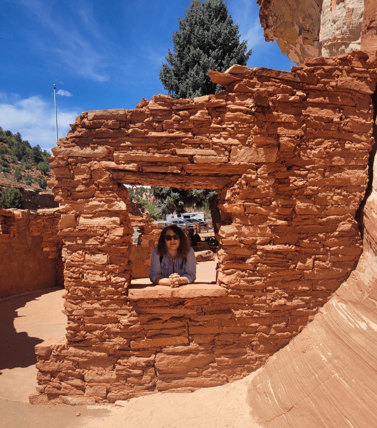 Person with sunglasses leaning on a brick window frame in an ancient ruin, blue sky above, trees in background.