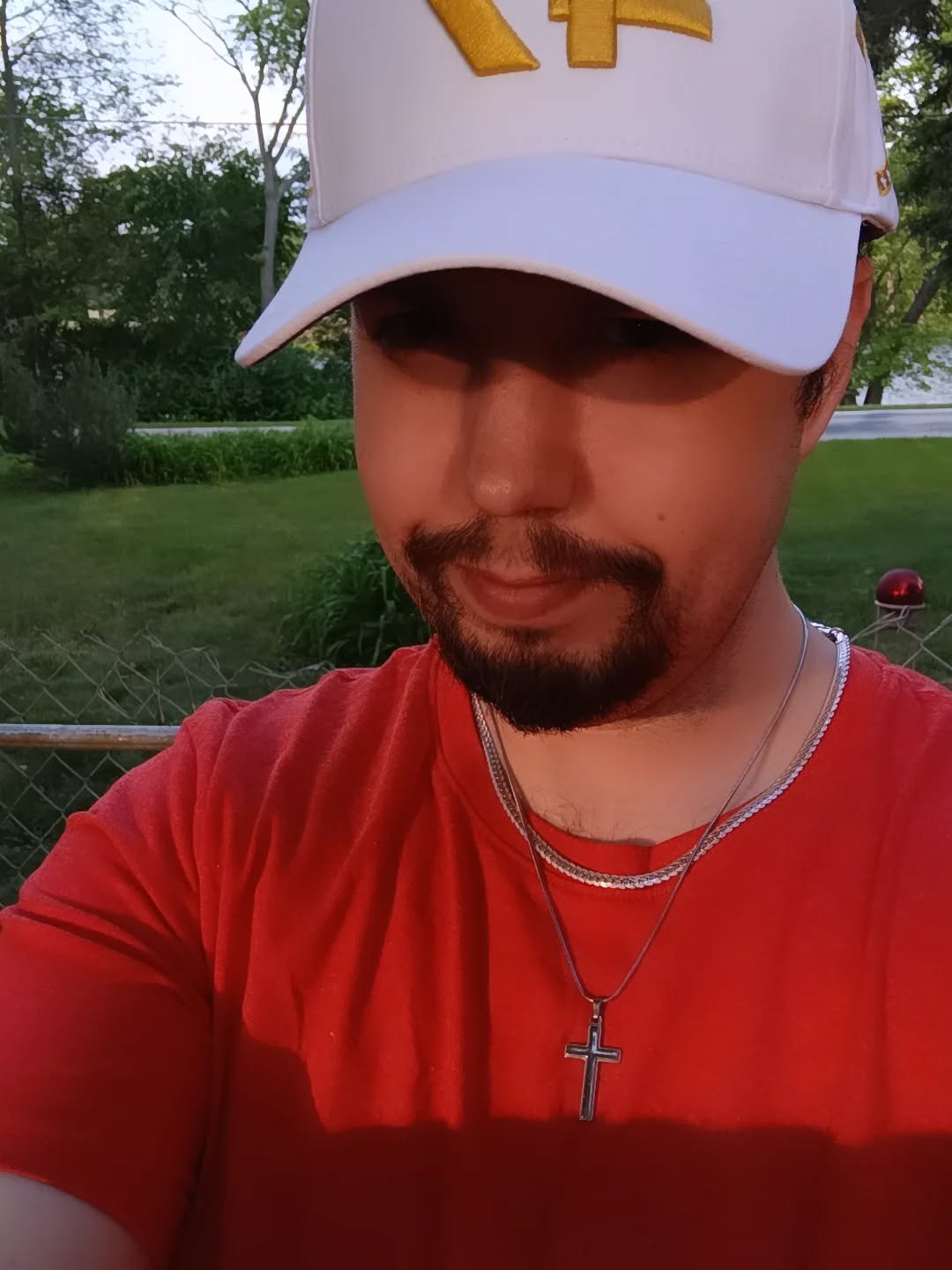 Person wearing a white cap with yellow design, red shirt, outdoors with trees and fence, smiling slightly.