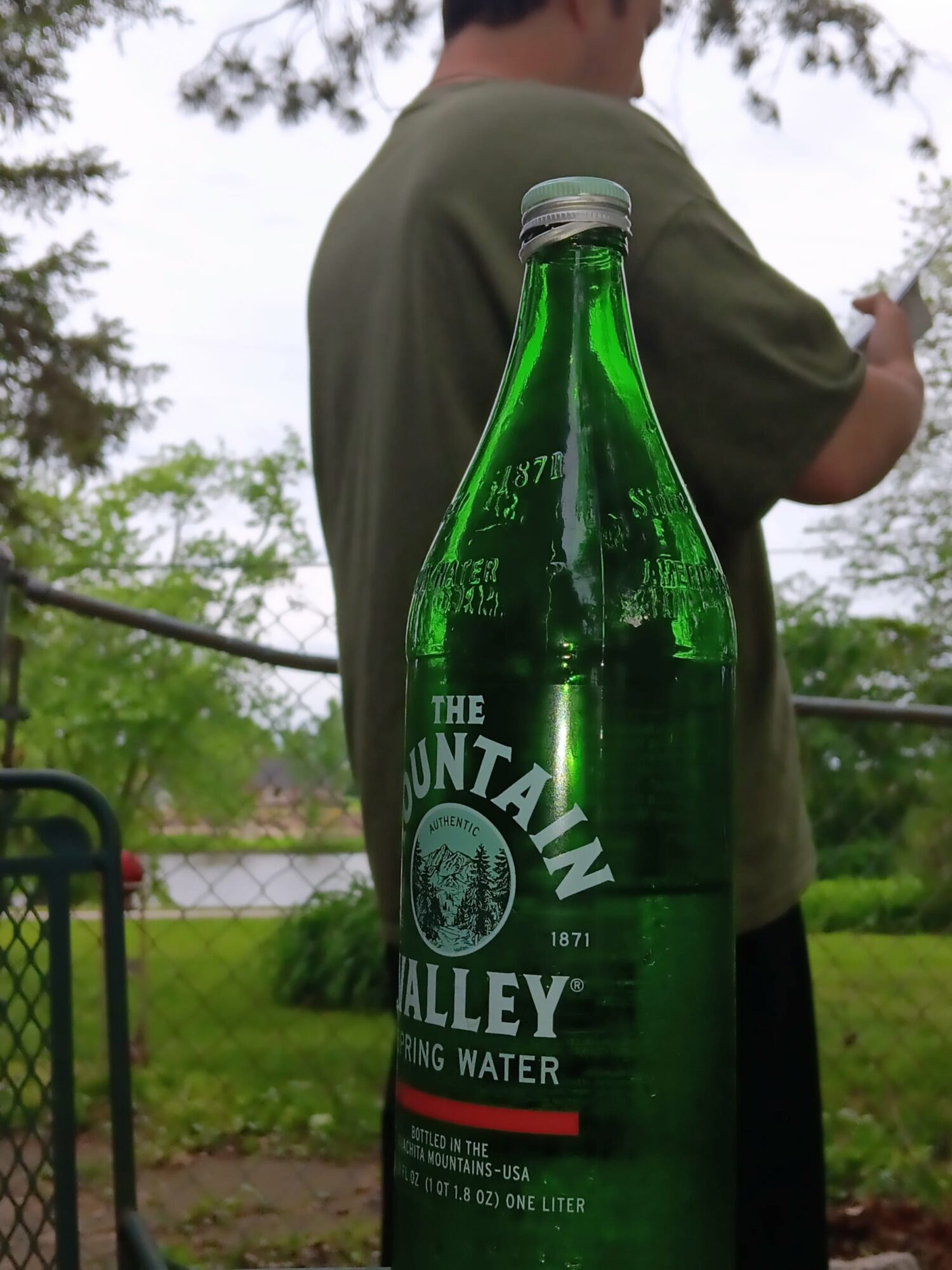 Person in green shirt outdoors with trees and fence, bottle of Mountain Valley spring water in foreground.