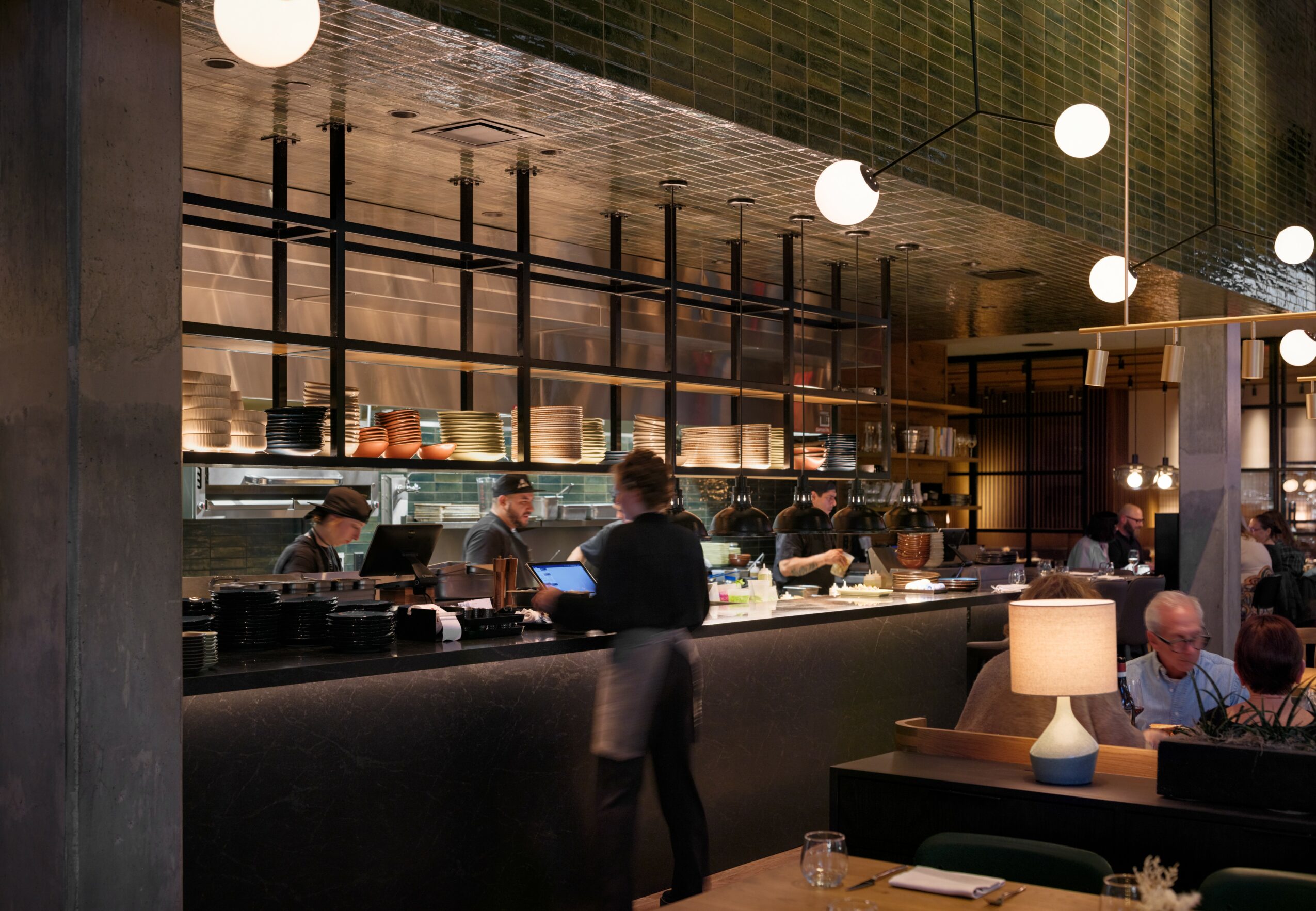 Interior of a modern restaurant or bar with a visible open kitchen, people working behind the counter, and patrons seated at tables.