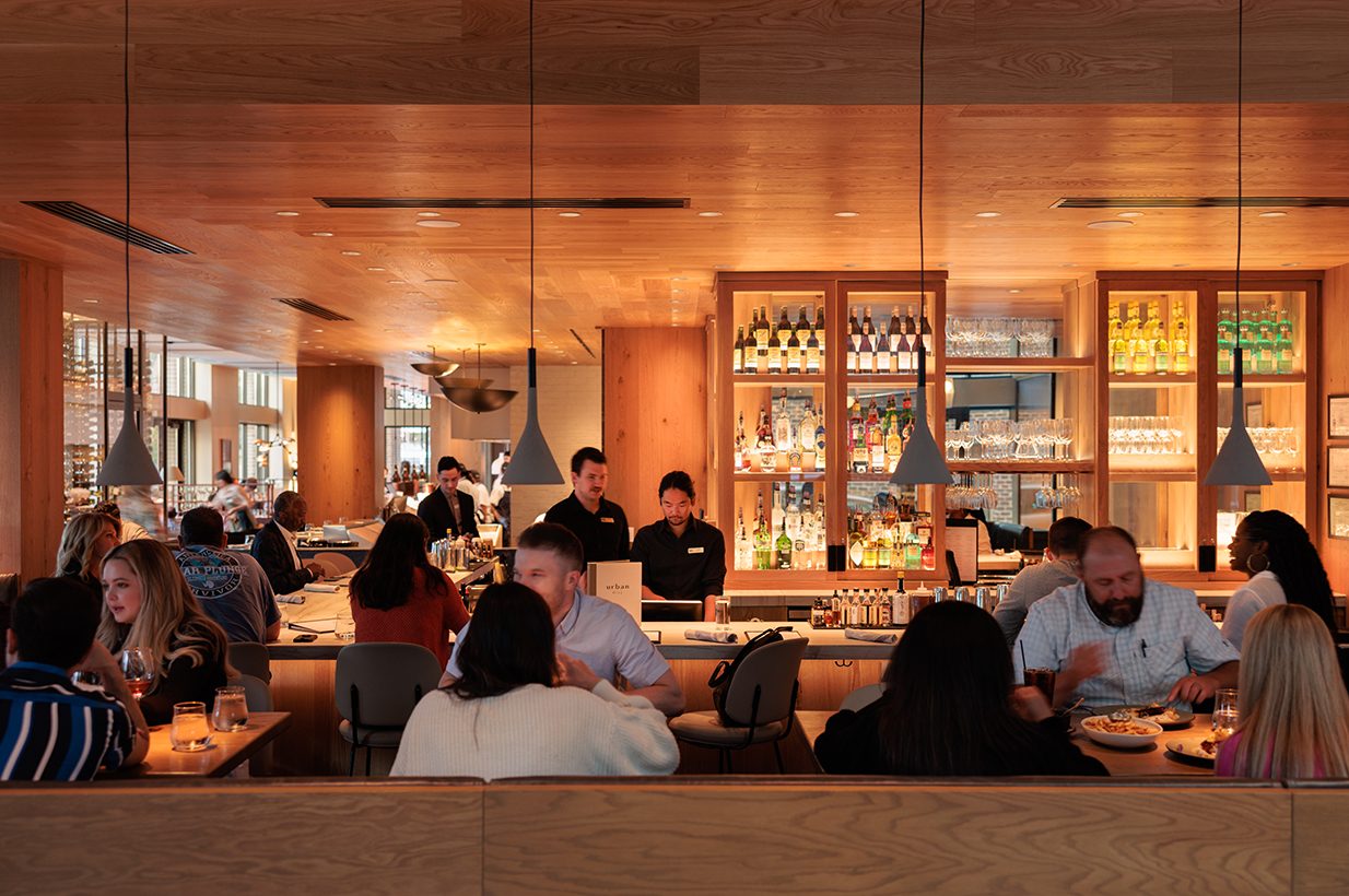 People dining in a restaurant with wooden walls and shelves displaying bottles, some engaged in conversation, others eating.