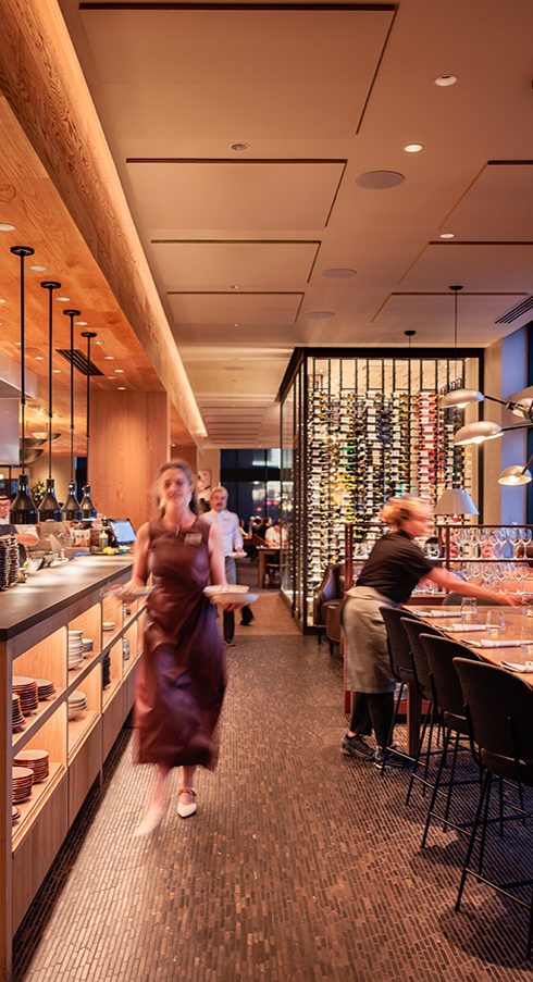 Woman in a maroon dress walking in a restaurant with bar seating and decorative partition, warm lighting, and wooden accents.