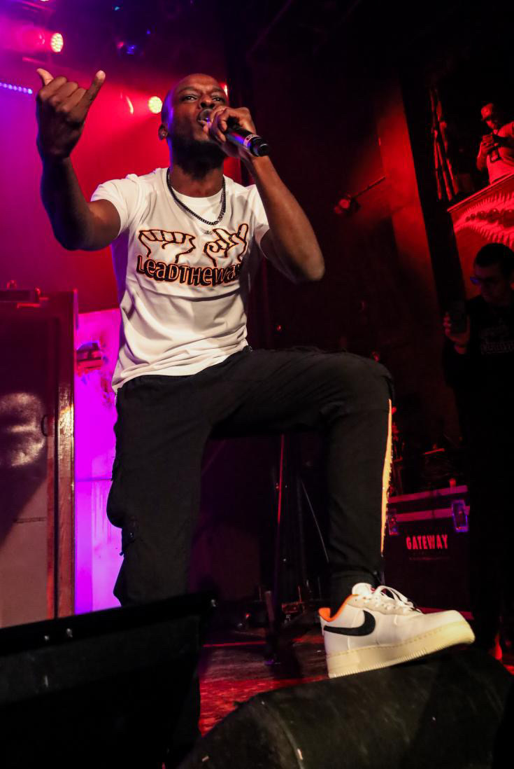 Performer singing into microphone on stage with colorful lighting, wearing a white T-shirt and black pants, one foot on a monitor.