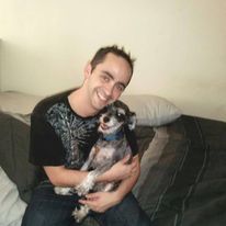Person smiling and holding a small dog on a couch in a room with plain wall