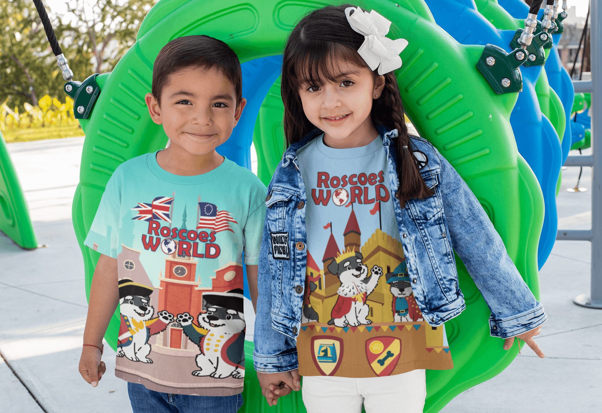 Two children stand in front of a green playground structure, smiling, wearing colorful T-shirts with cartoon characters, outdoors.