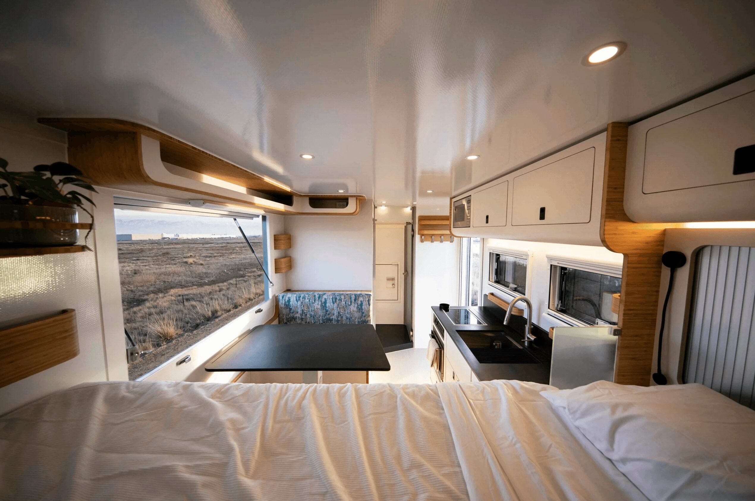 Interior of a camper with a bed, window, and kitchen area, showing a view outside landscape, ceiling lights, and storage cabinets.