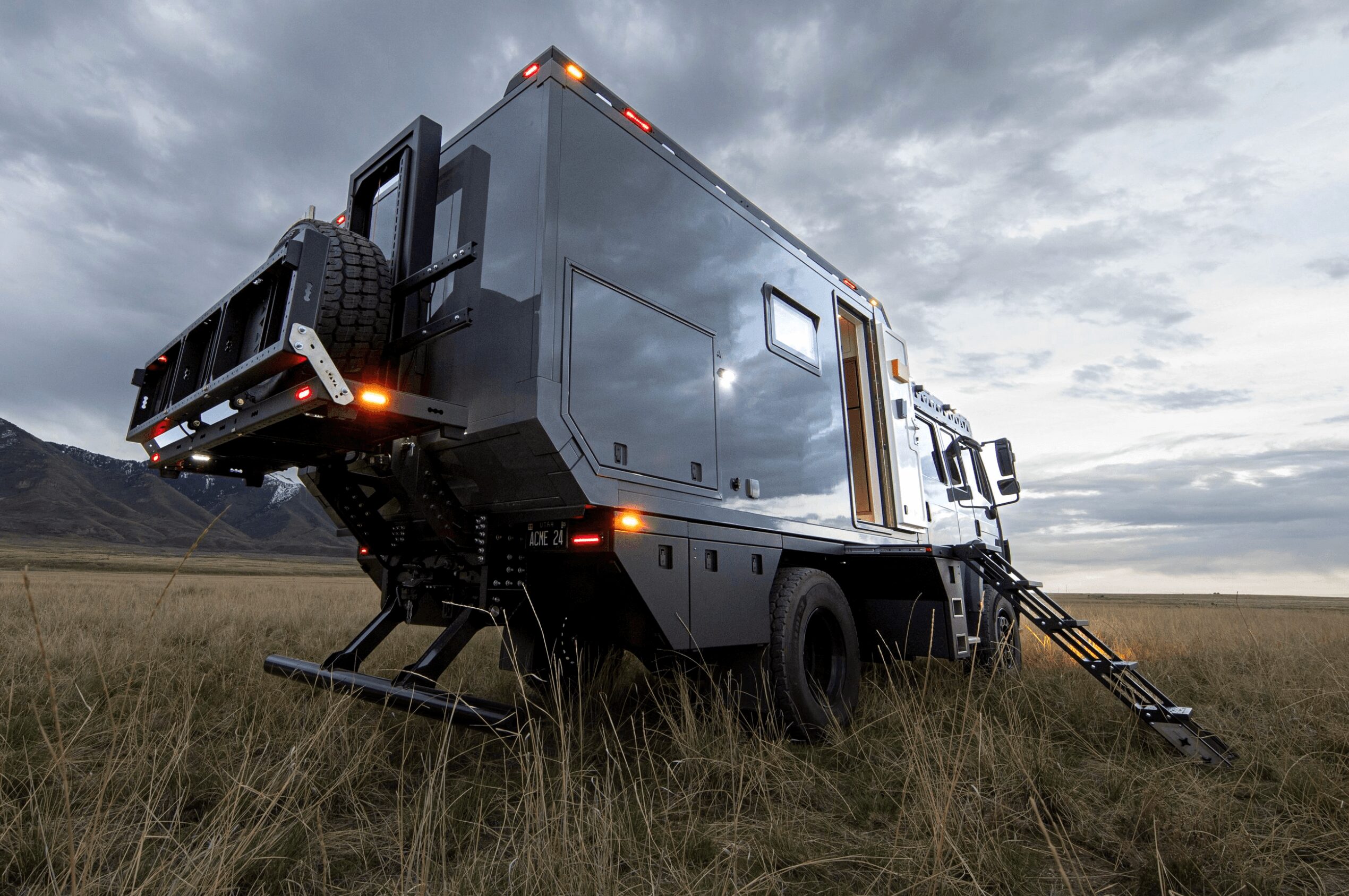 Large black off-grid camper vehicle parked in grassy field with mountains and cloudy sky in background.