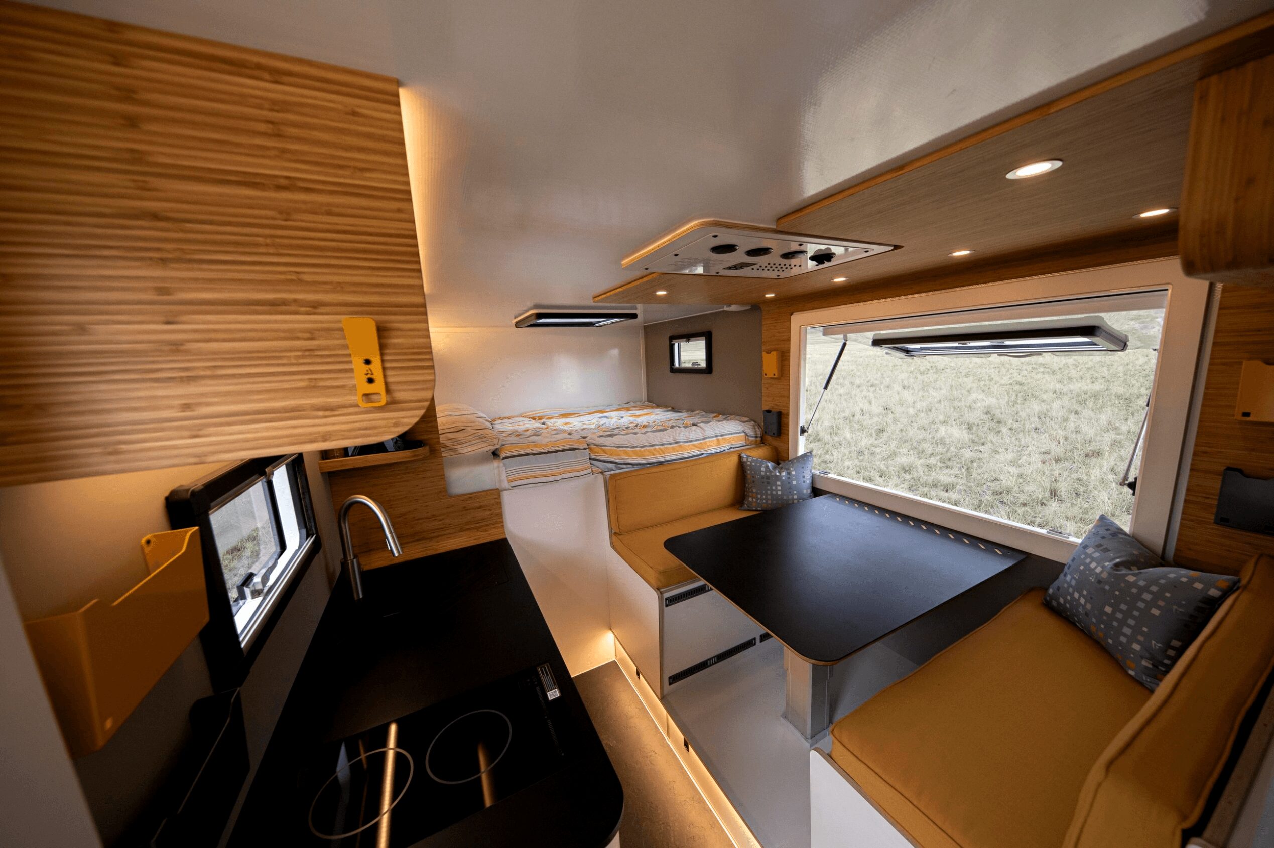 Interior of a camper van with a black table, window, and seating area with cushions, wood paneling, and a stove.