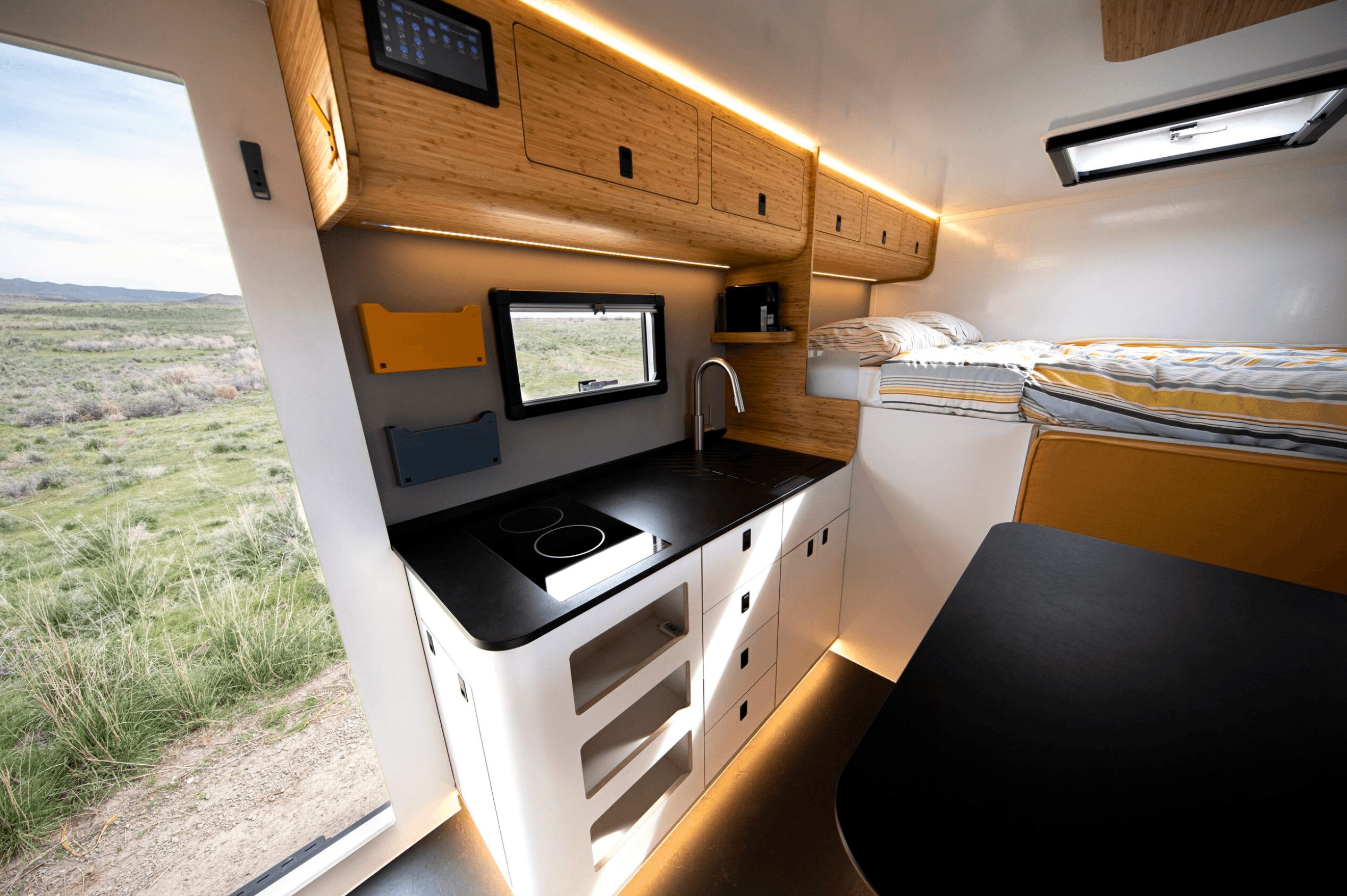 Compact kitchen area with black countertop, sink, and stove inside a camper van, with a bed nearby and a window showing outdoor scenery.