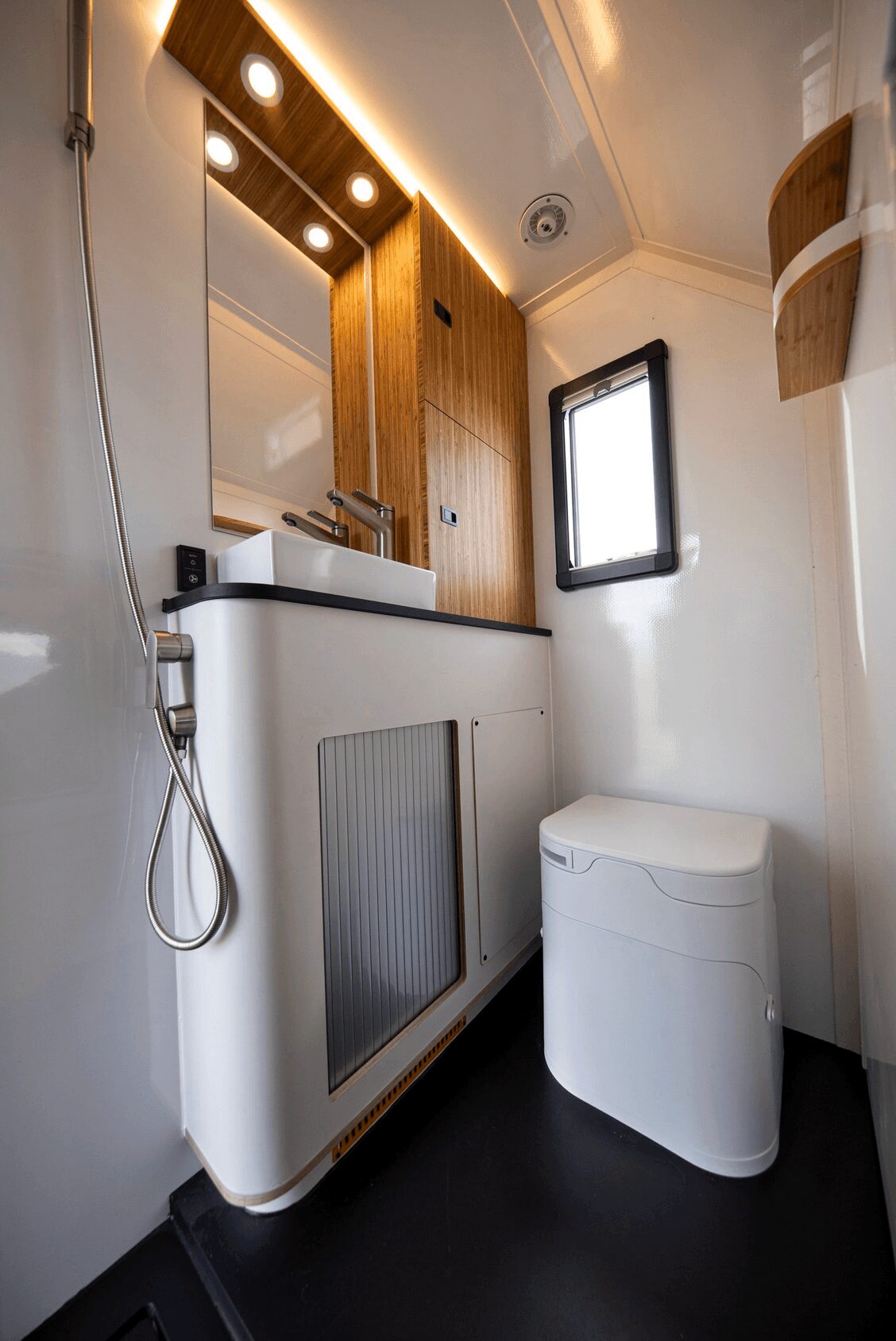 Compact bathroom with a mirror, wooden cabinet, window, and a white toilet, with a black floor and ceiling lighting.