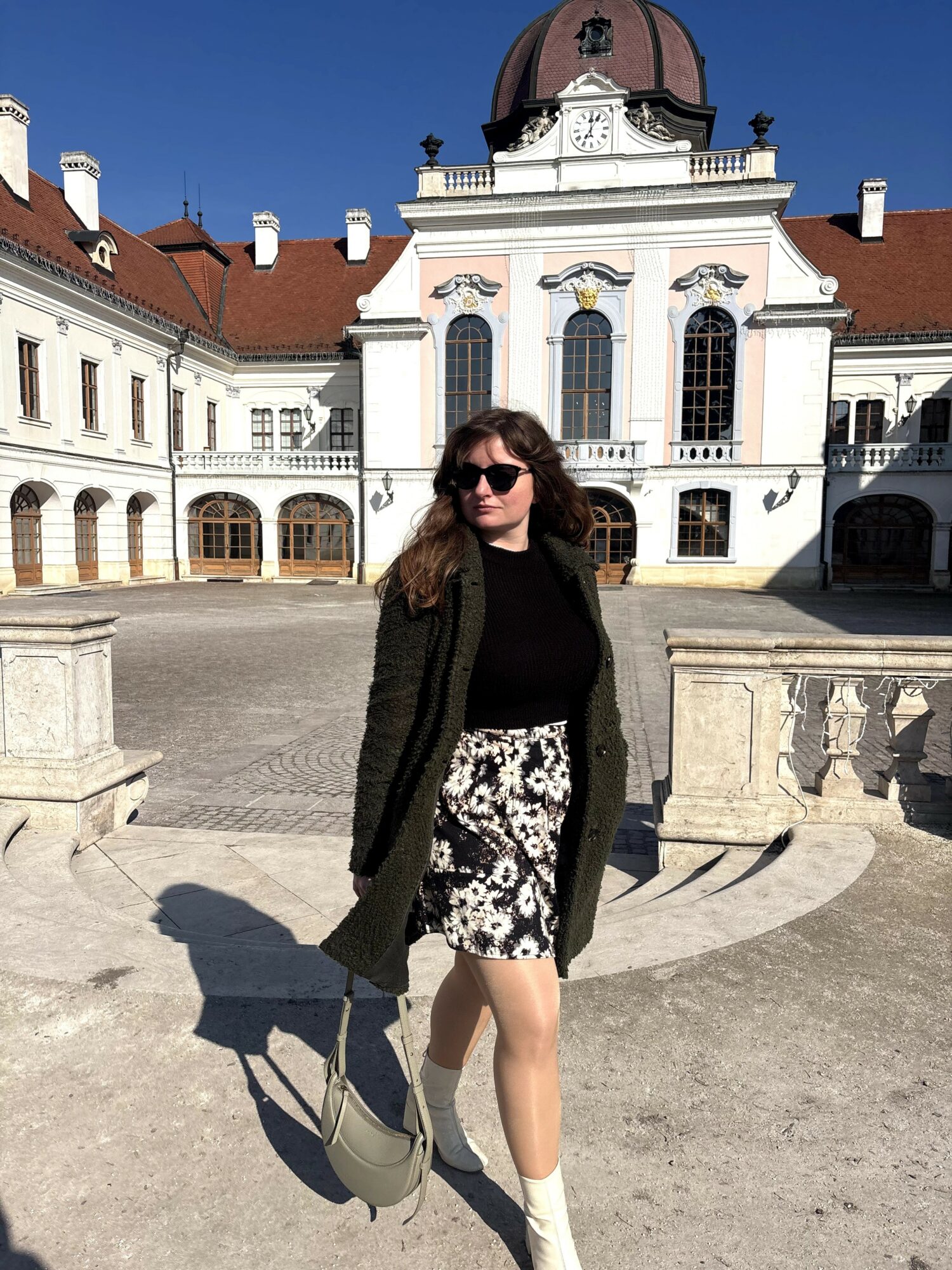 Woman with sunglasses standing outdoors in front of a historic building with a domed roof, sunny sky, and stone balustrades.