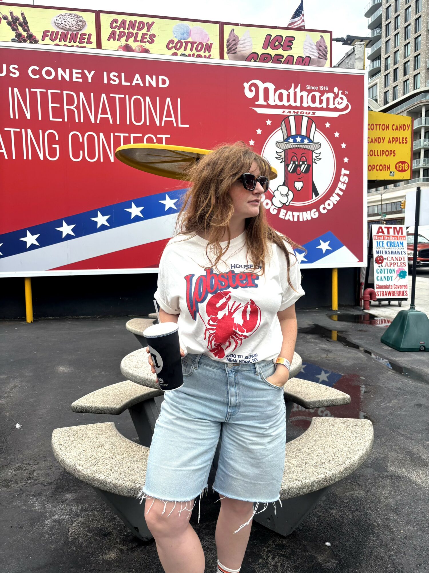 Woman with sunglasses and curly hair stands in front of a red and white sign, holding a drink, wearing a white t-shirt and denim shorts.
