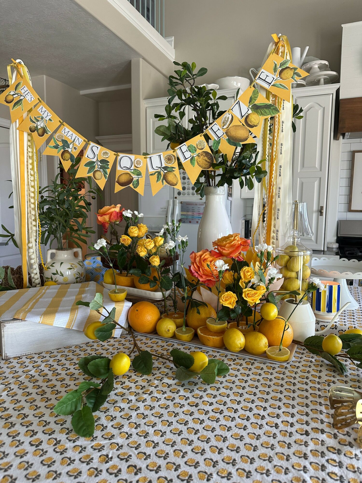 Decorative table with yellow and orange flowers, lemons, and greenery, arranged with candles and a patterned tablecloth.