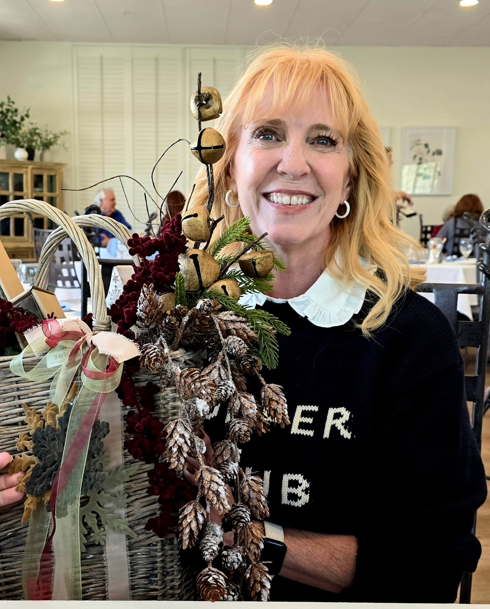 Smiling woman with blonde hair holding a decorated floral arrangement in a room with tables and people.